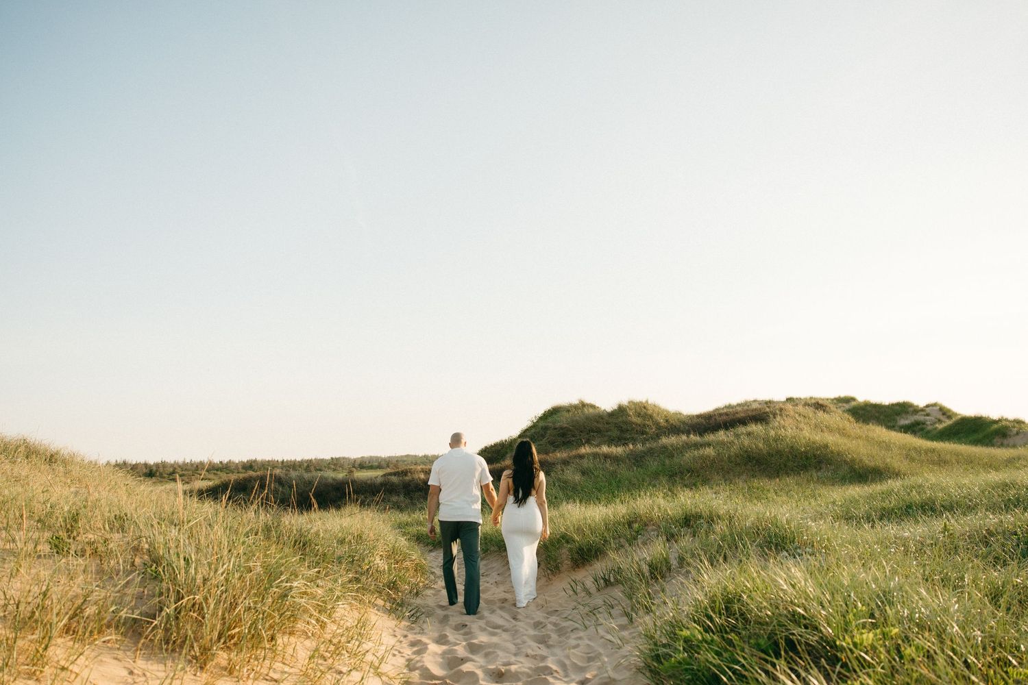 Couple holding hands walking along a sandy beach path surrounded by green dunes on a clear sunny day.