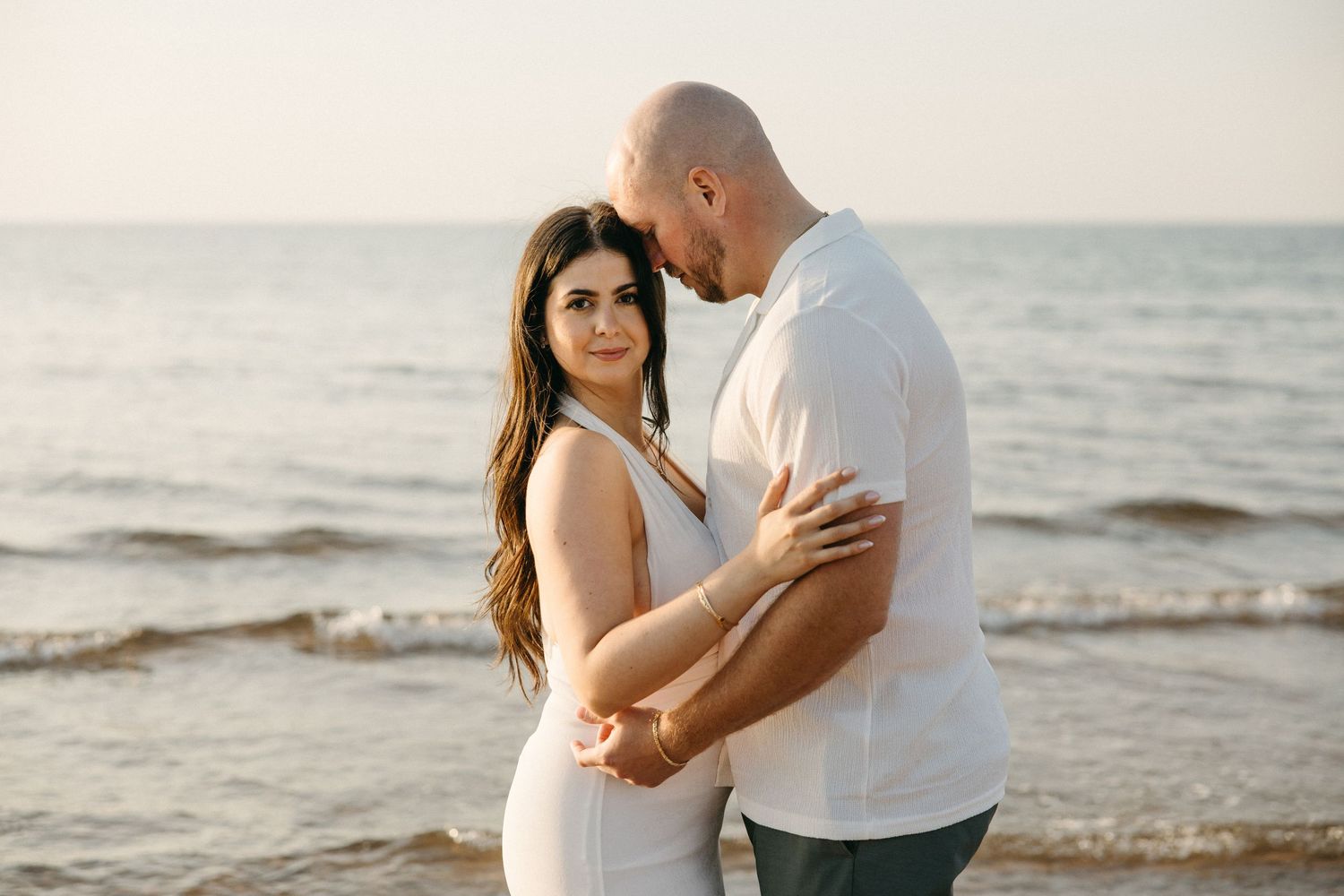 Couple embracing on beach shoreline at golden hour, woman in white halter dress looking at camera.