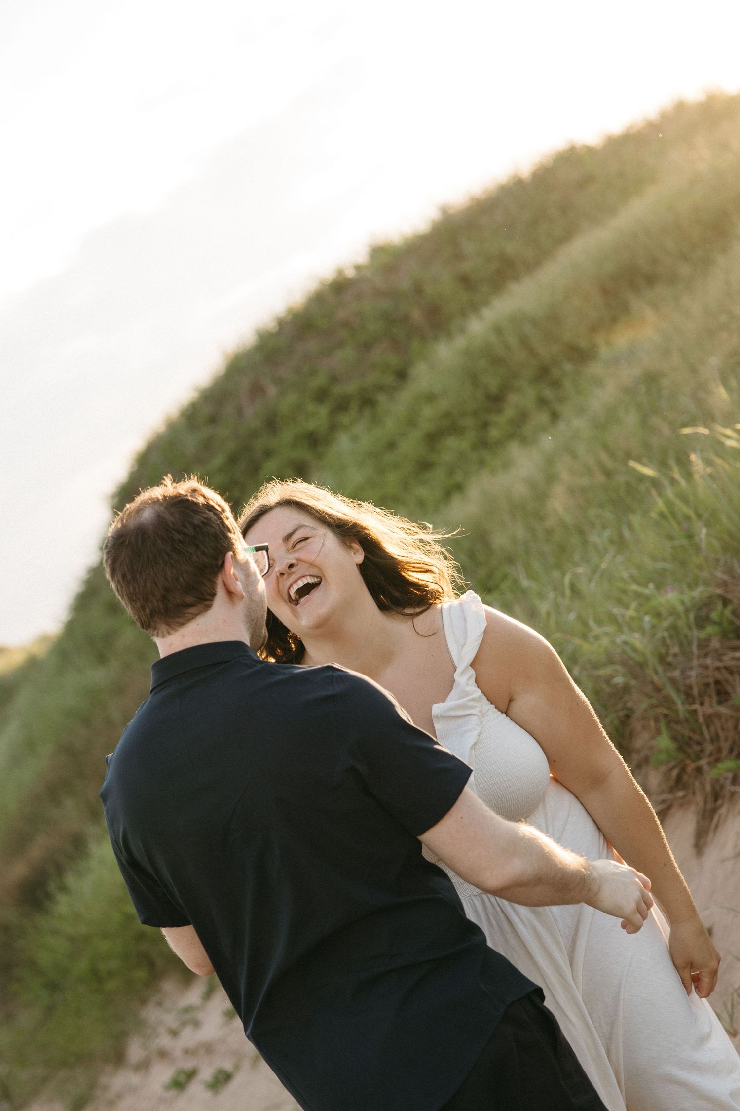Couple laughing and dancing together on sandy beach dunes at golden sunset.