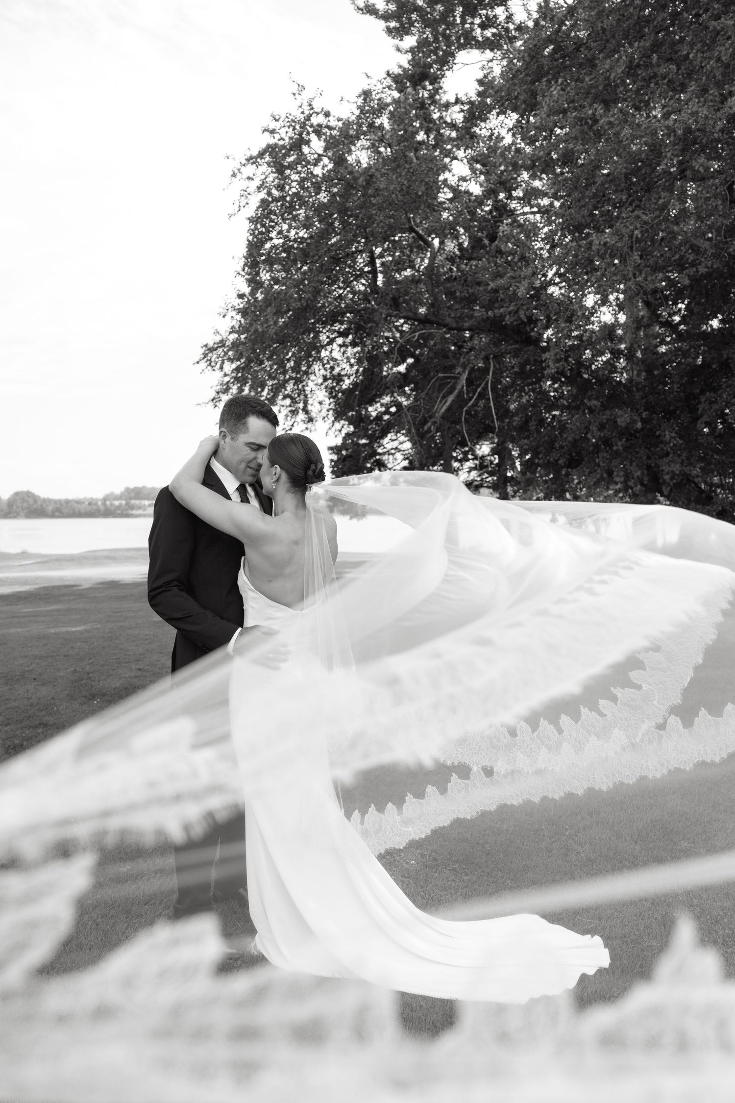 Black and white wedding photo of bride and groom embracing outdoors, veil flowing in wind, trees and water in background.