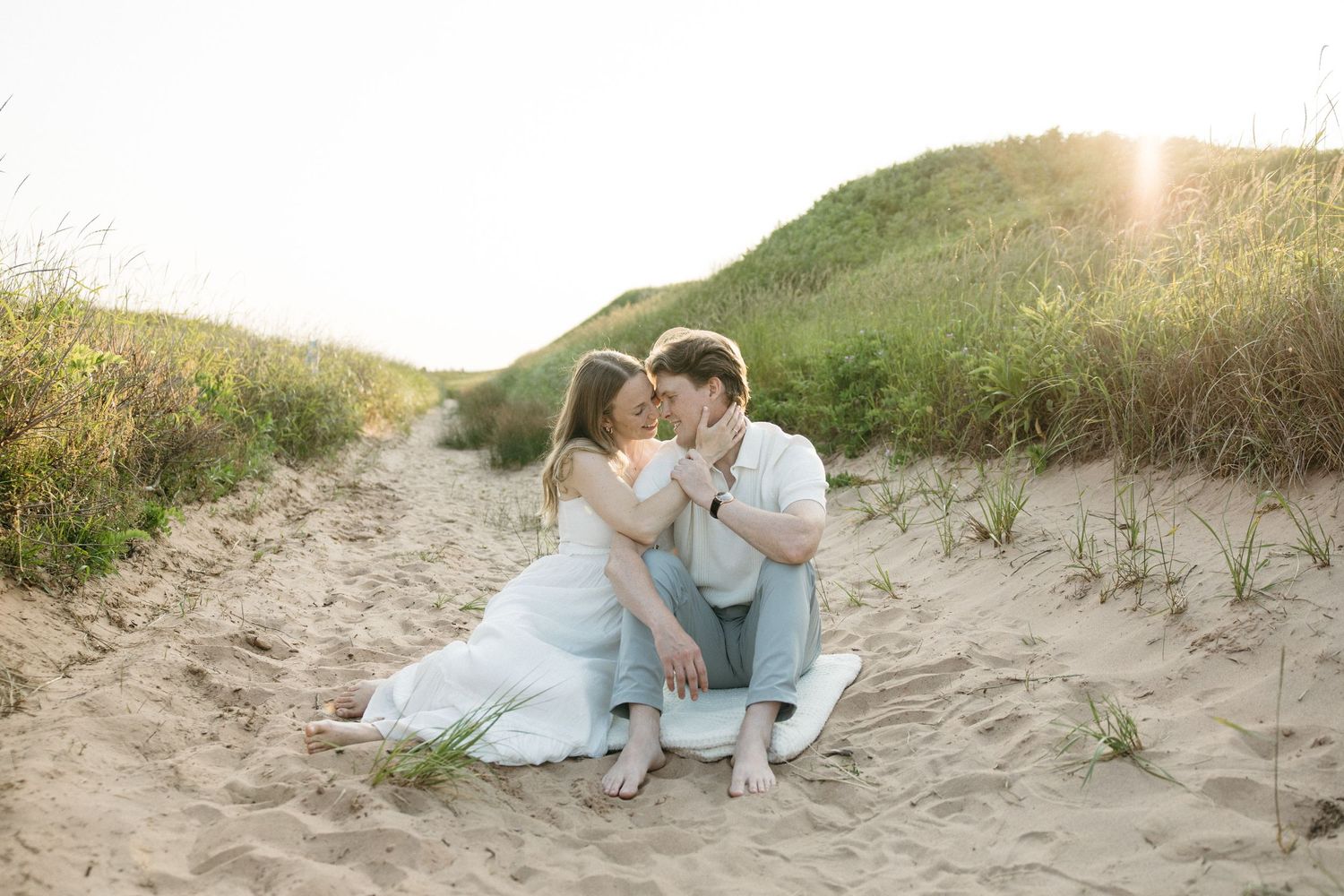 Engaged couple sitting together on sandy beach path surrounded by dunes at sunset.
