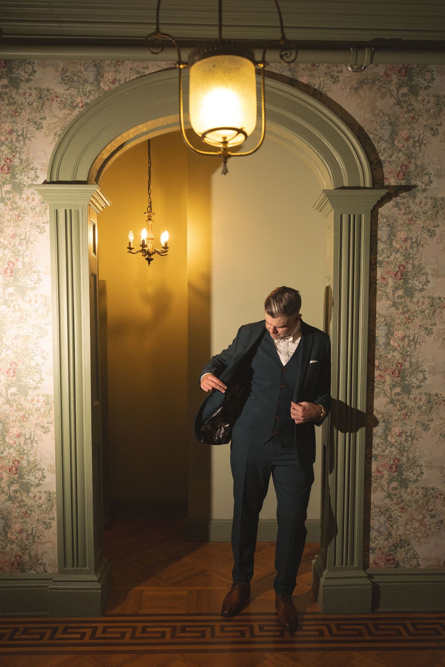 Groom in a three-piece navy suit adjusts his jacket while standing in an arched doorway with warm vintage lighting.