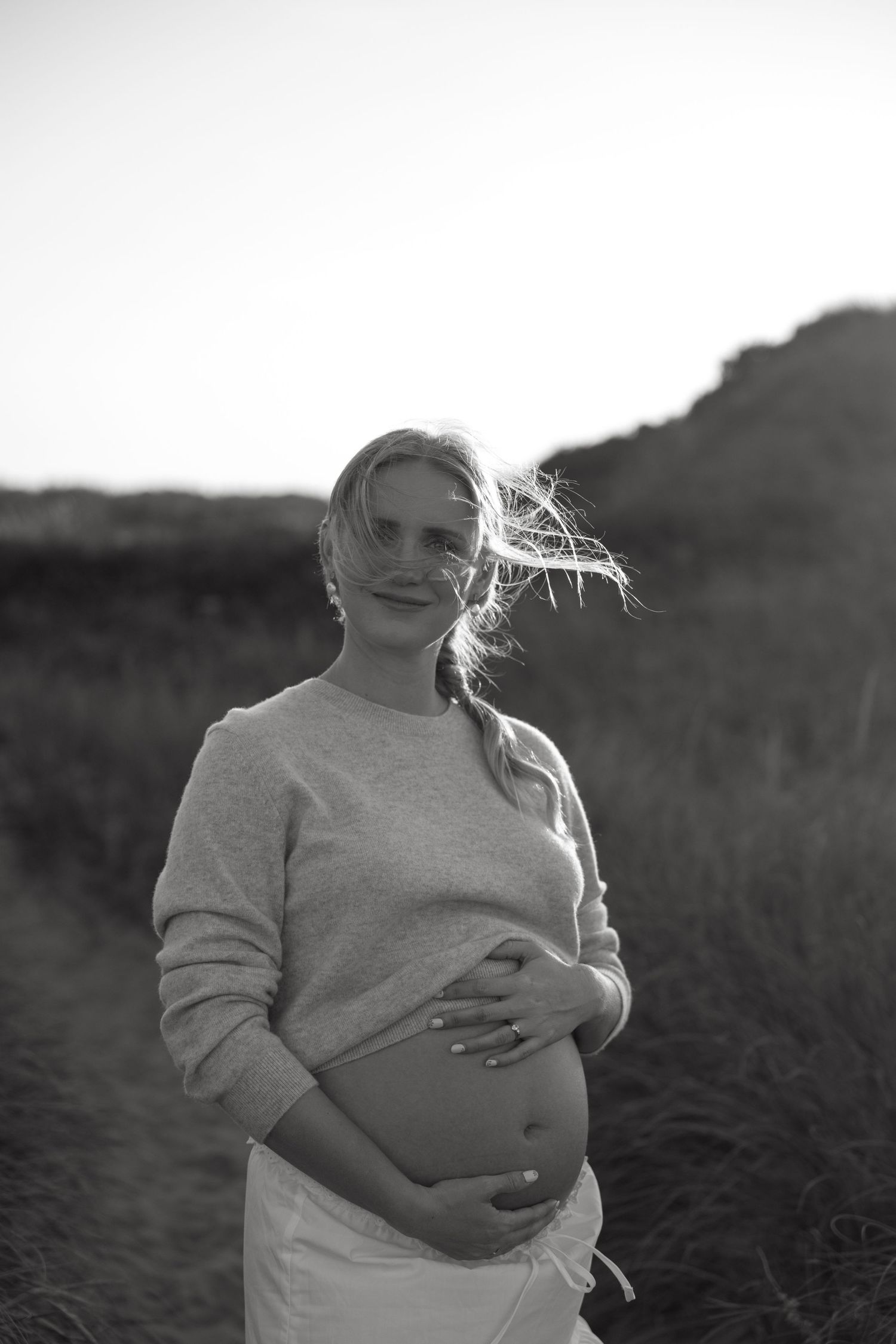 Black and white portrait of expectant mother with windswept hair in natural setting.