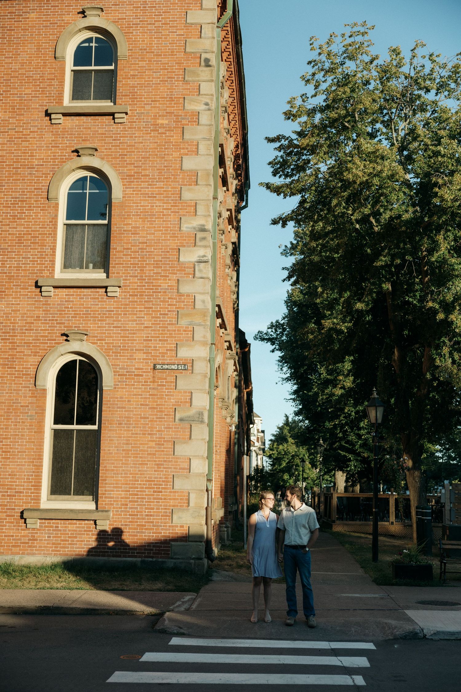 Two figures stand near a brick building with arched windows at sunset.