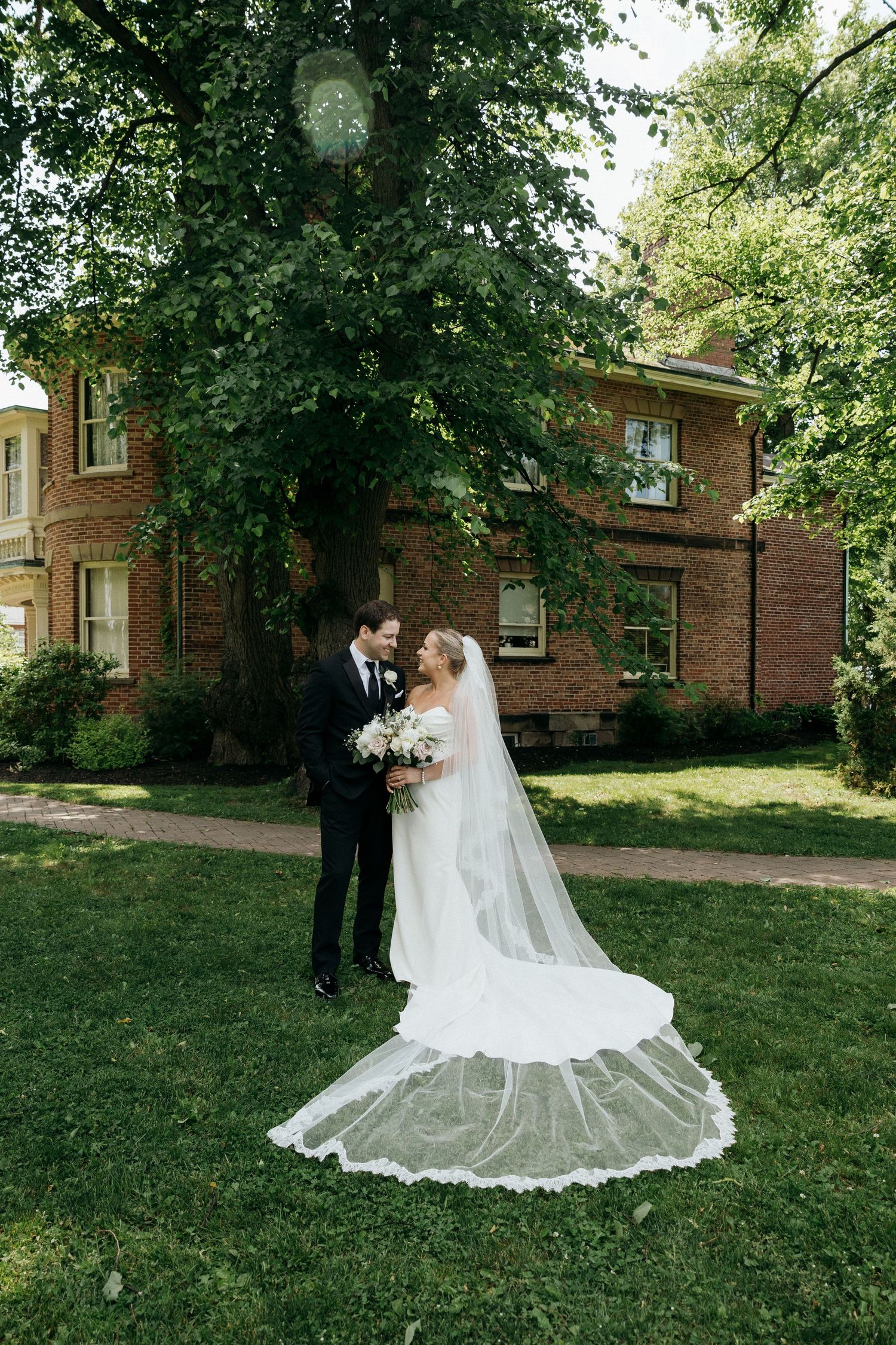A wedding couple poses on a grassy lawn in front of a classic brick building on their special day.