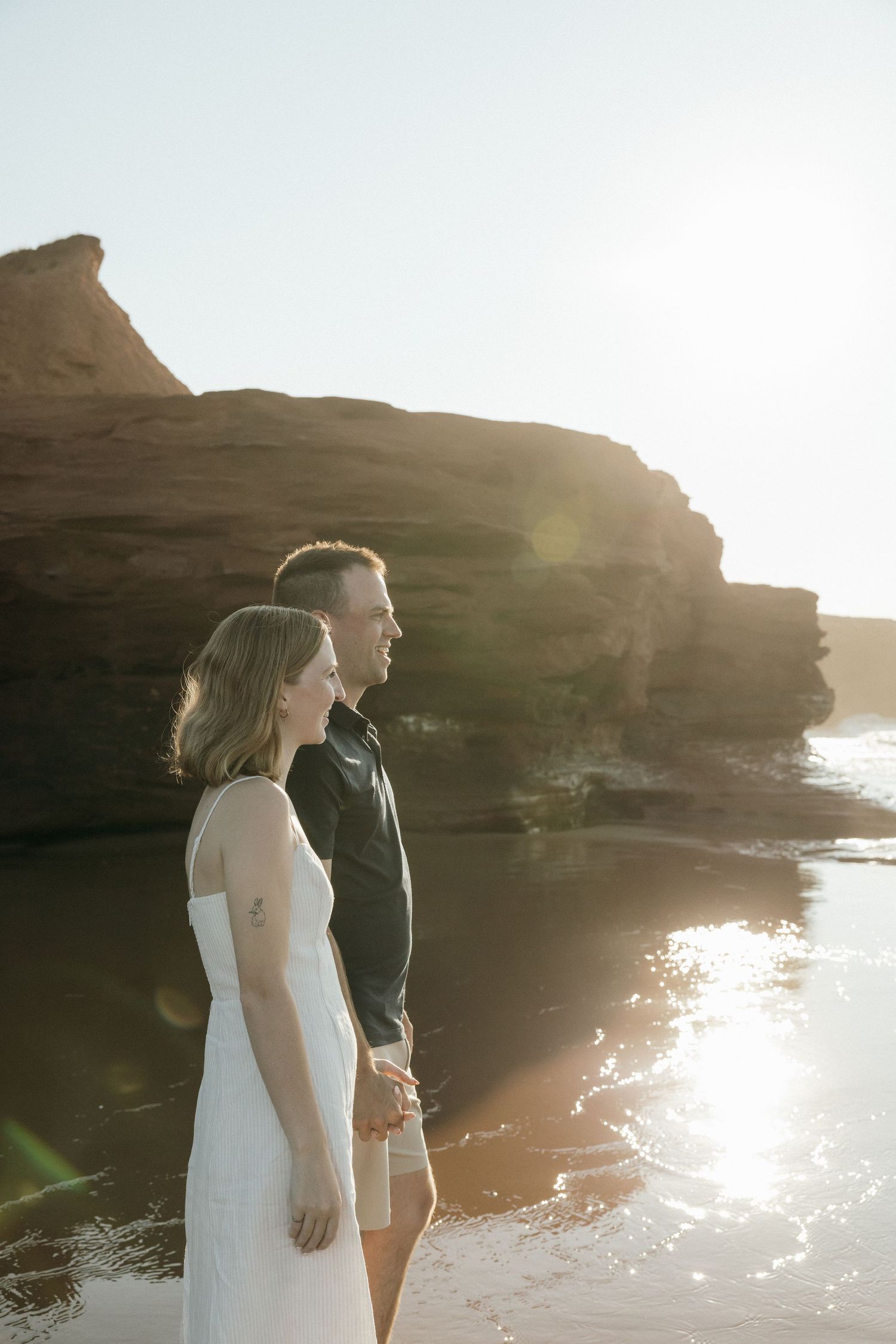A couple stands together overlooking the ocean from dramatic coastal cliffs at sunset.