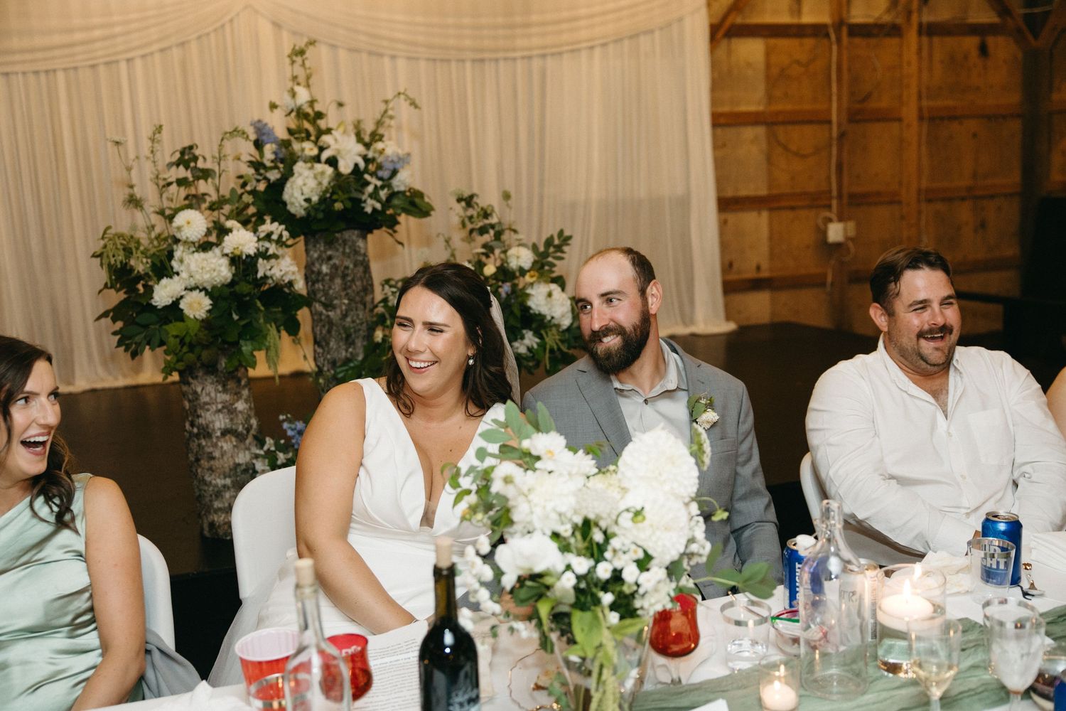 Elegant wedding reception table settings with white floral centerpieces and string lights in barn venue.