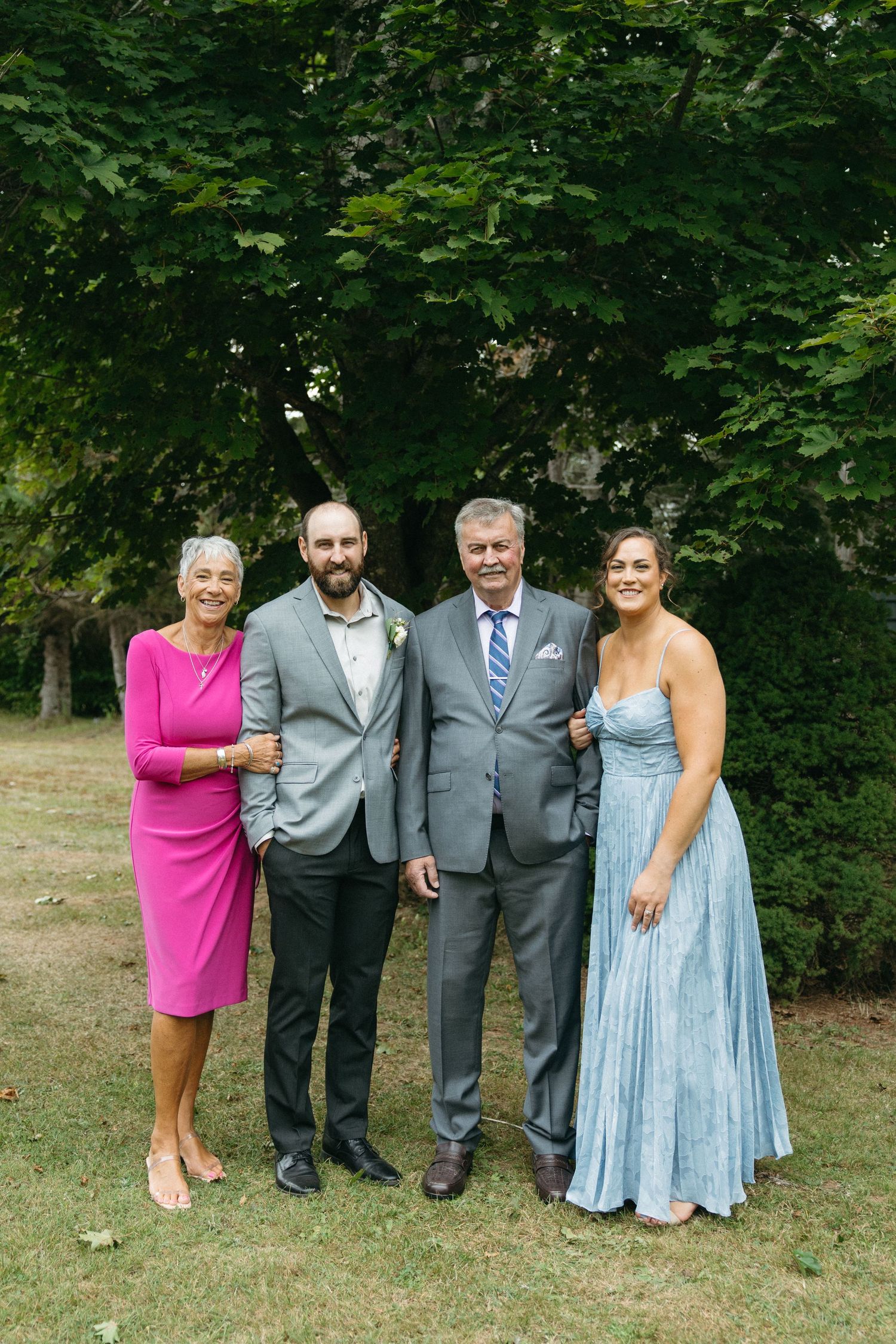 Wedding party in formal attire poses together on grassy lawn with trees in background.