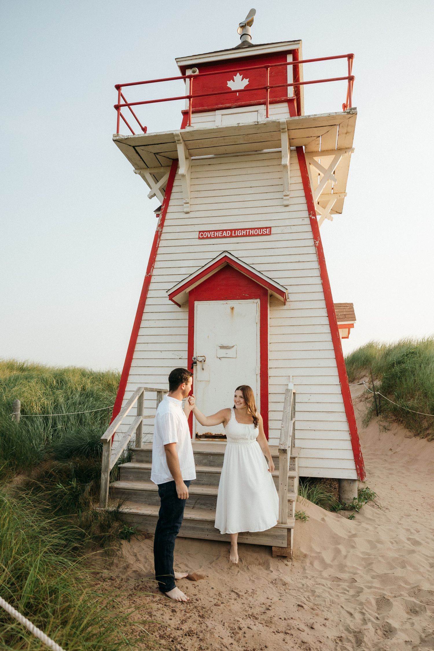 A couple stands on the steps of a red and white lighthouse during a romantic photoshoot.