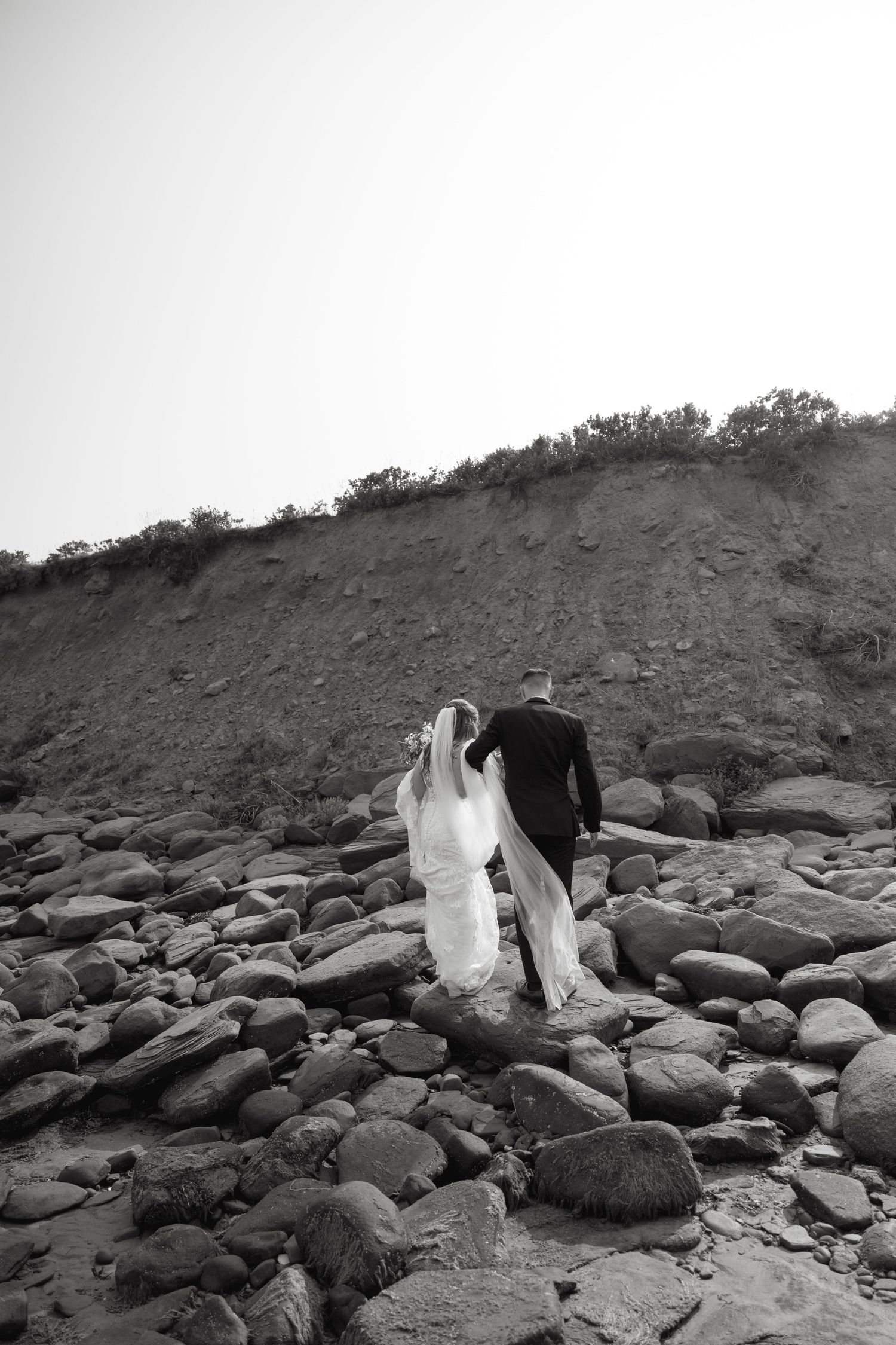 Black and white wedding photo on rocky beach with dramatic coastal backdrop.