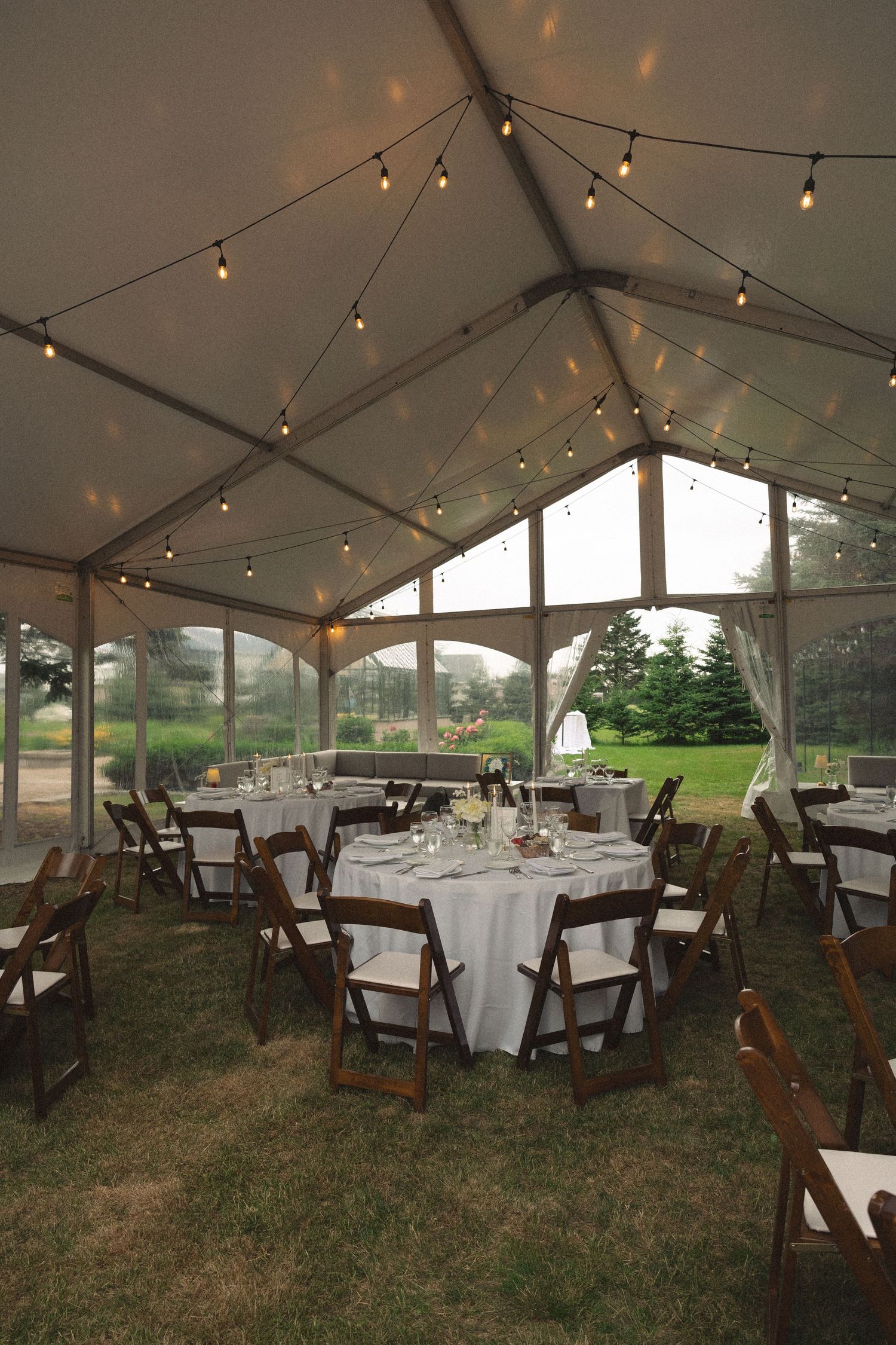 Elegant outdoor wedding reception tent with string lights, round tables, and wooden folding chairs.