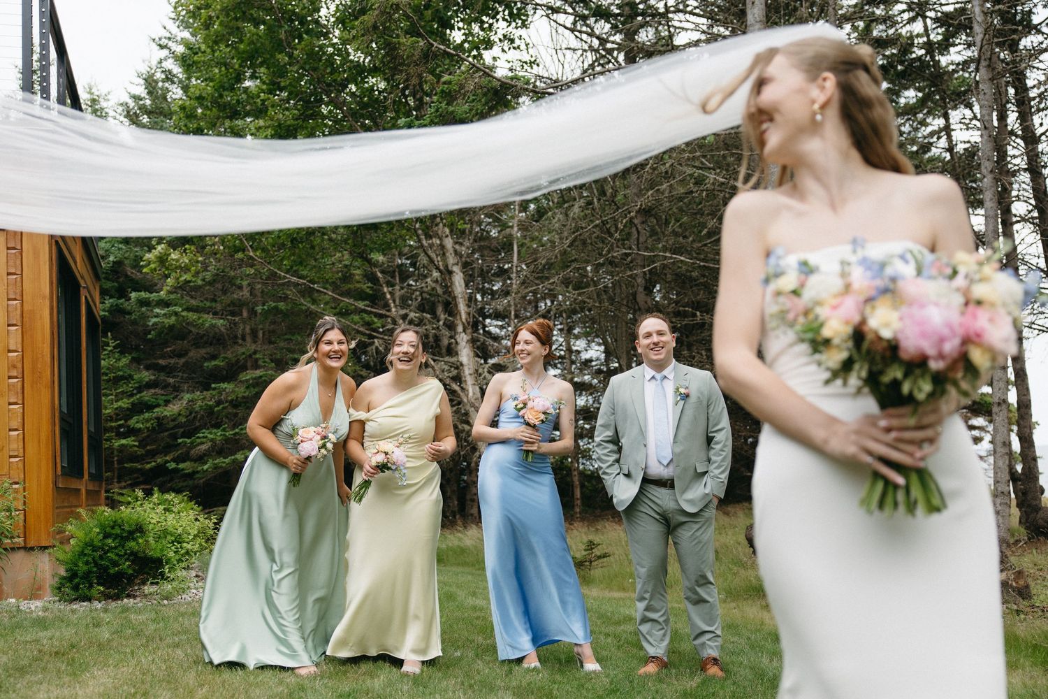 Bridesmaids in pastel gowns laugh joyfully as the bride's veil flows in the wind during outdoor wedding photos.