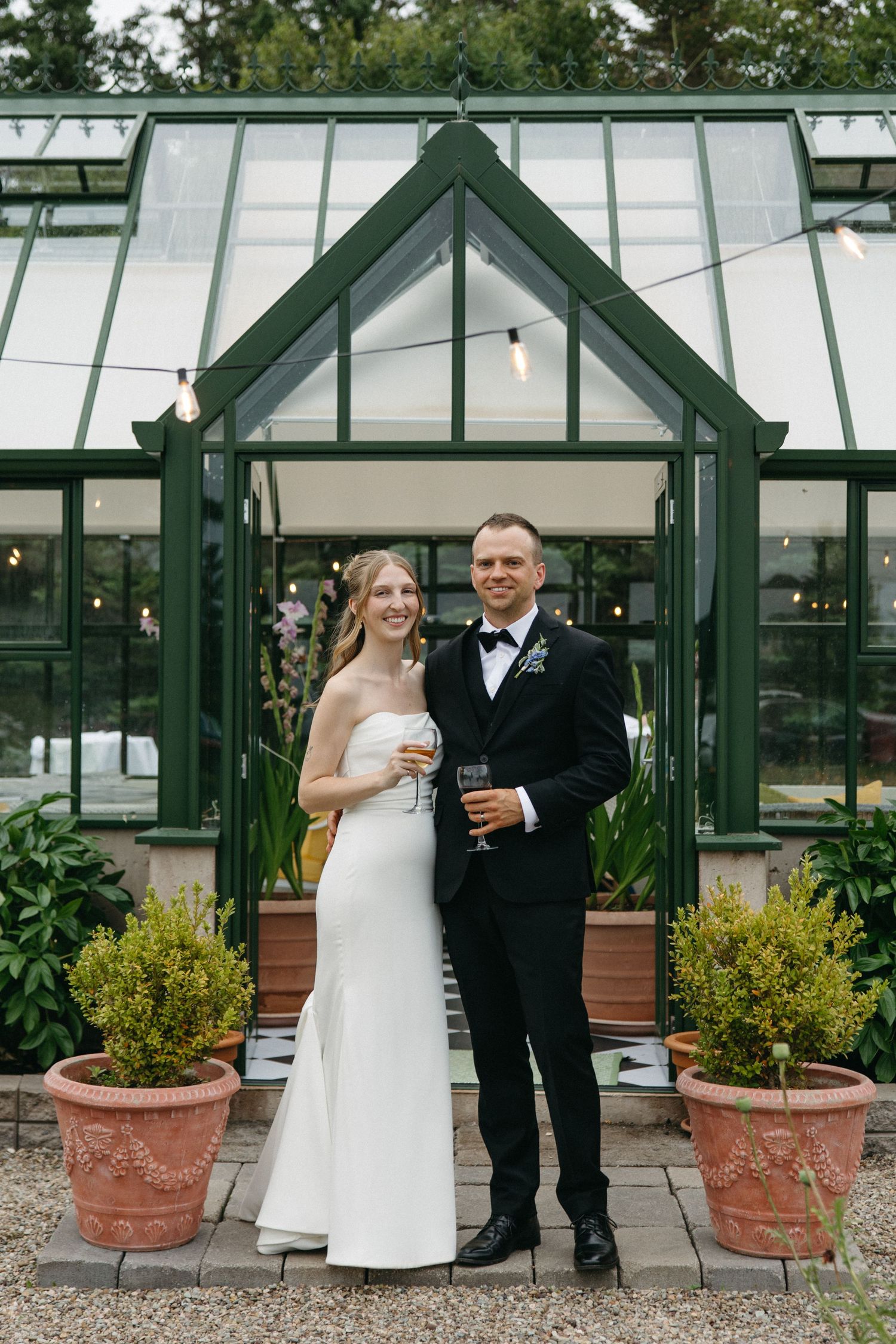 Smiling bride and groom holding drinks in front of elegant greenhouse on wedding day.