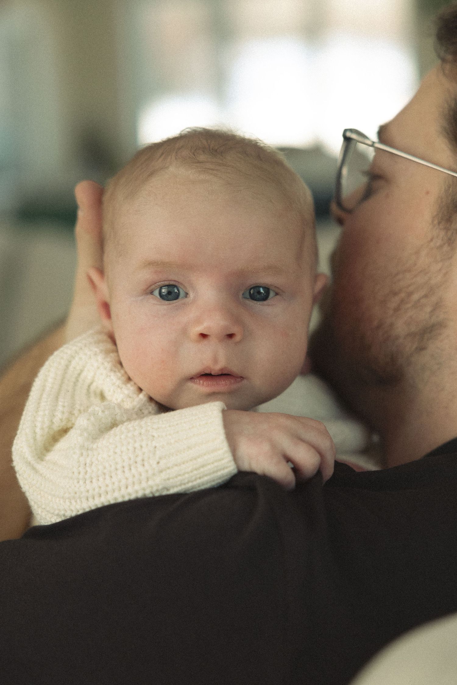 A baby in a cream sweater rests against someone's shoulder while looking at the camera with bright blue eyes.