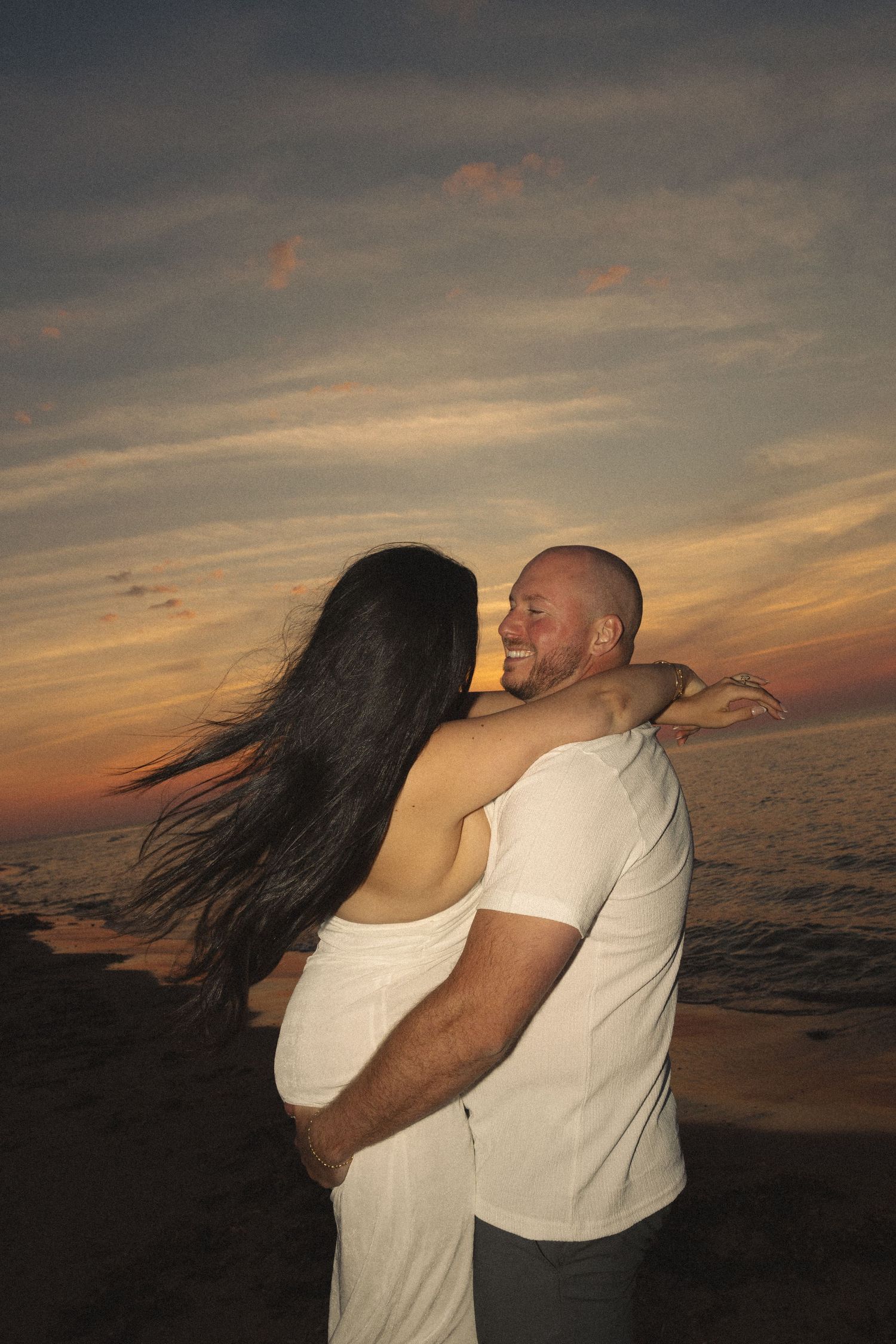 Happy couple hugging and laughing on beach at dramatic sunset, woman in backless white dress, ocean in background.