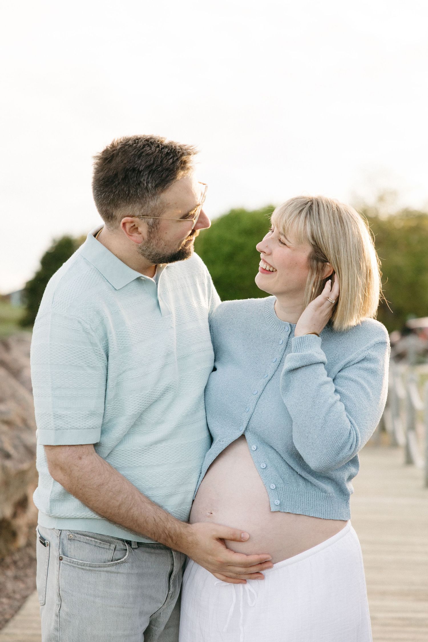 Smiling pregnant couple embrace tenderly on a boardwalk, man holding woman's baby bump in golden light.