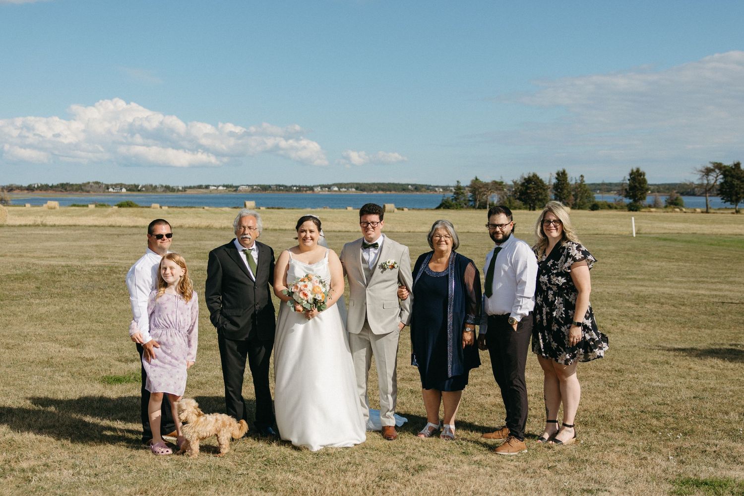 Wedding party family portrait on green lawn with scenic lake and blue sky in the background.