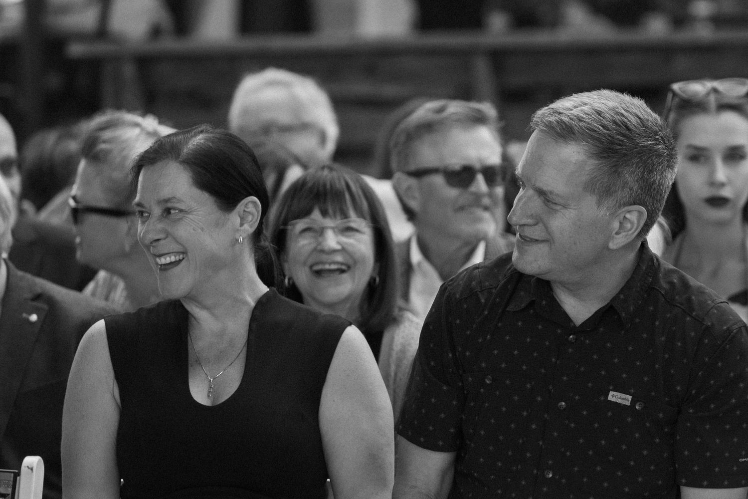 Black and white candid photo of smiling wedding guests enjoying an outdoor ceremony in a crowd.
