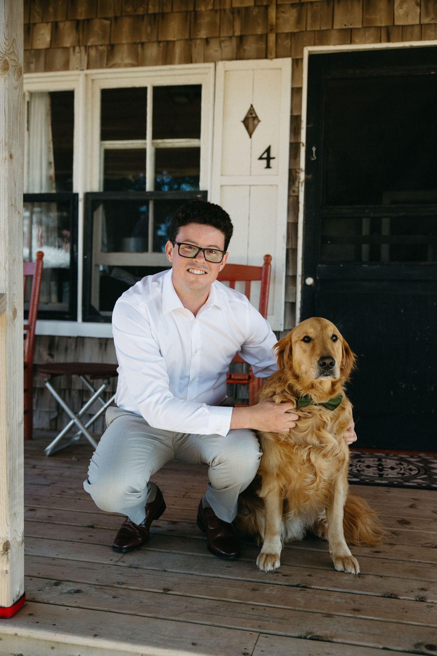 Smiling groom in white shirt and gray pants poses with golden retriever on rustic cabin porch.