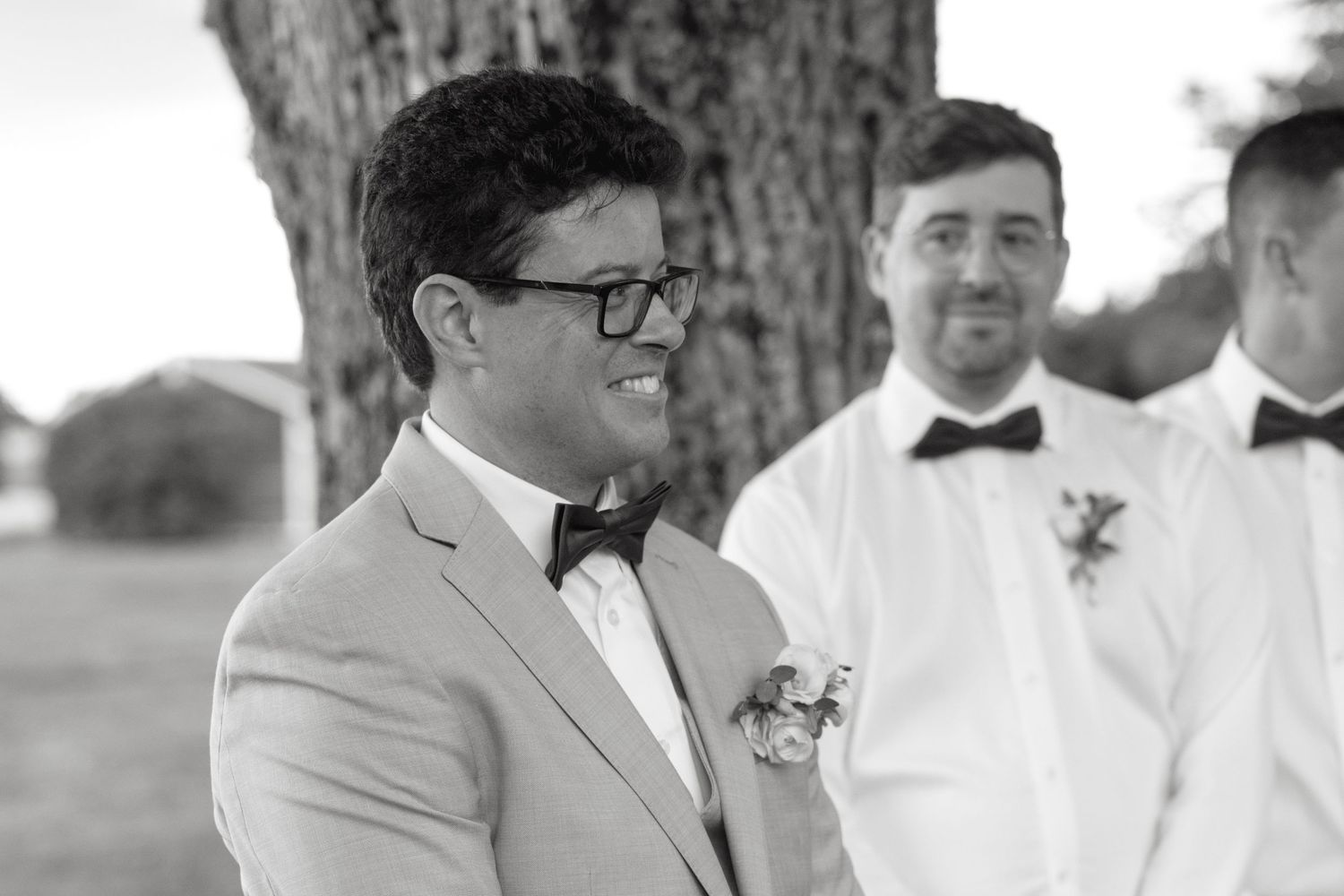 Black and white photo of smiling groom in bow tie standing by tree with groomsman in background.
