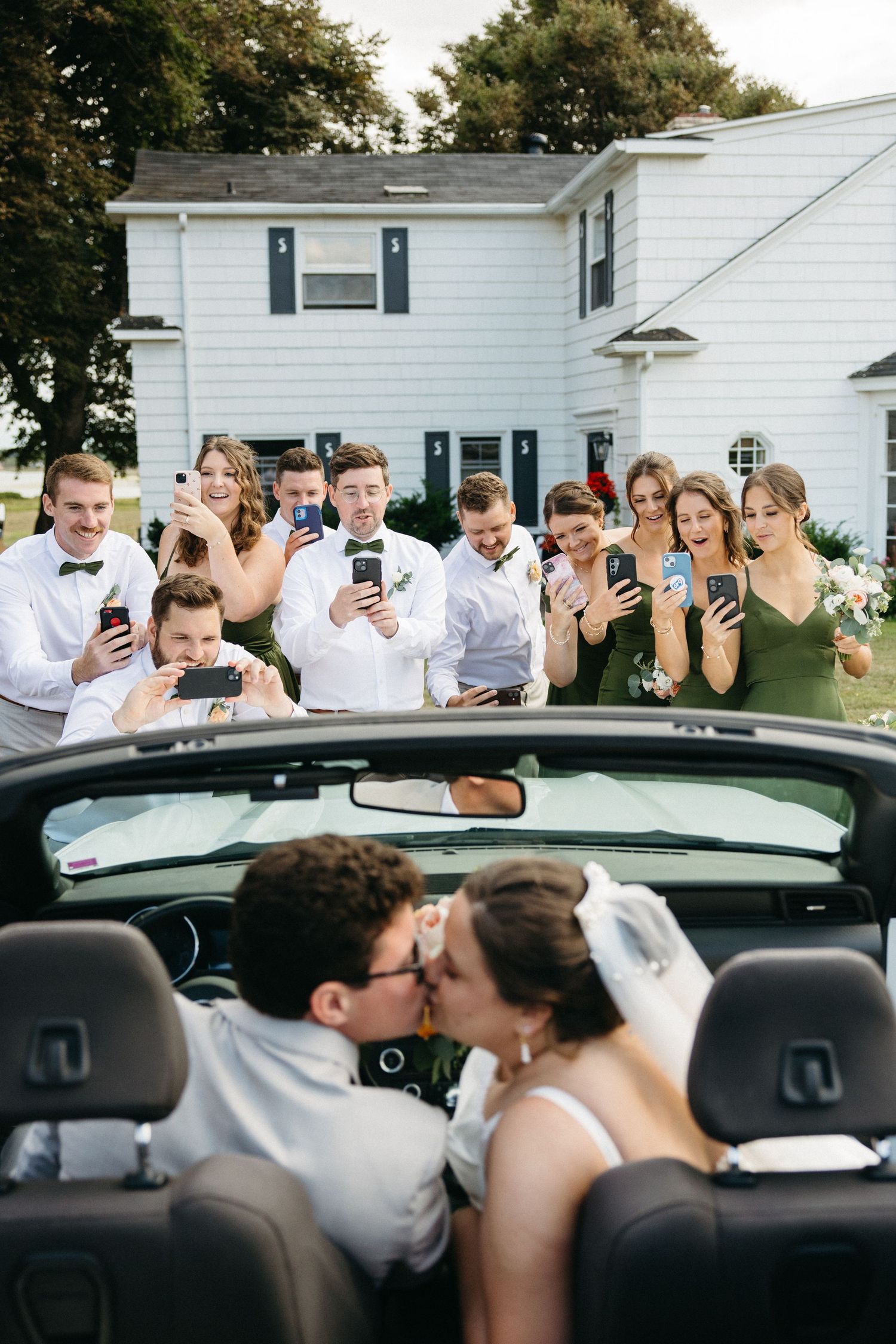 Bridal party photographing bride and groom kissing in a convertible car after wedding ceremony.
