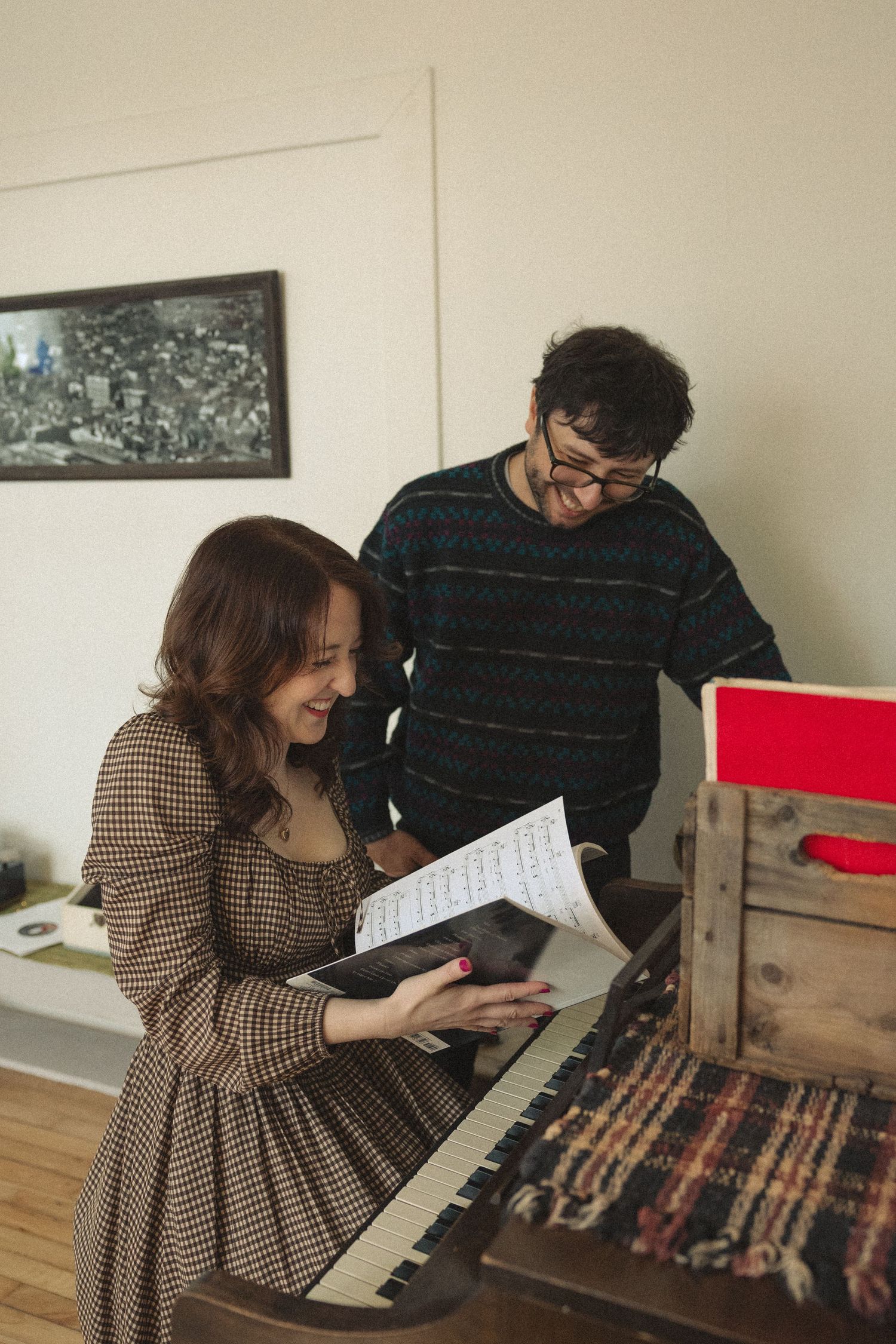 A young woman smiles at sheet music at a piano while a man in glasses looks on in a cozy room.