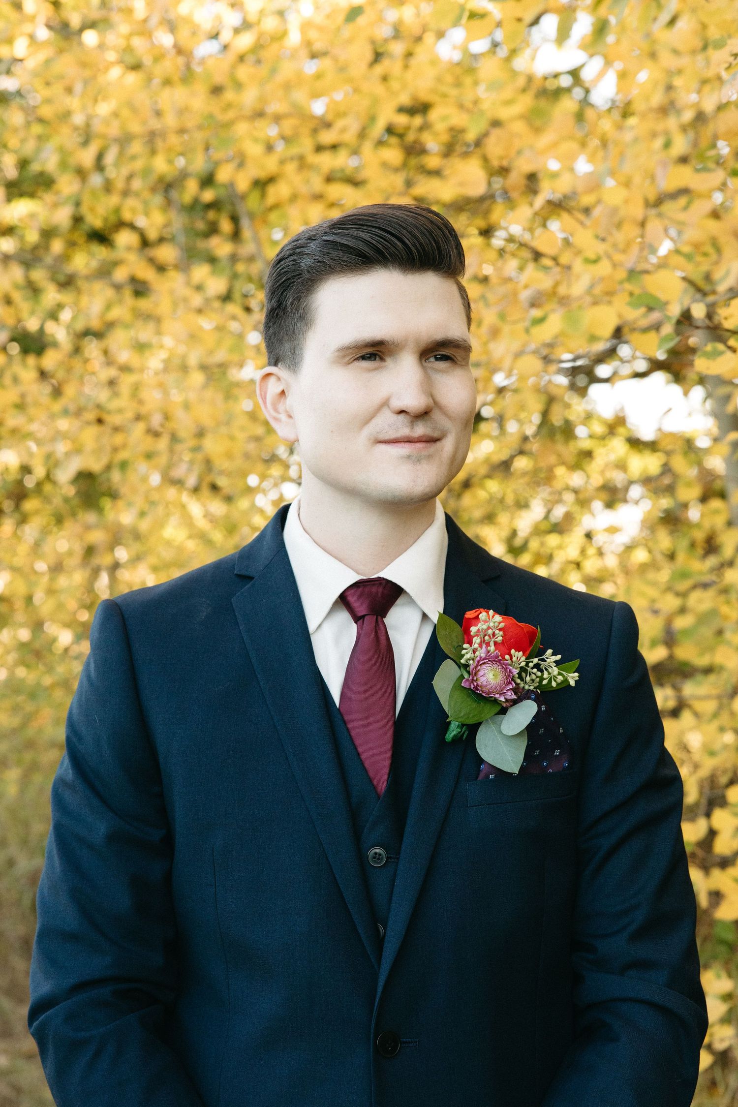Groom in navy suit with burgundy tie and red boutonniere poses against fall foliage.