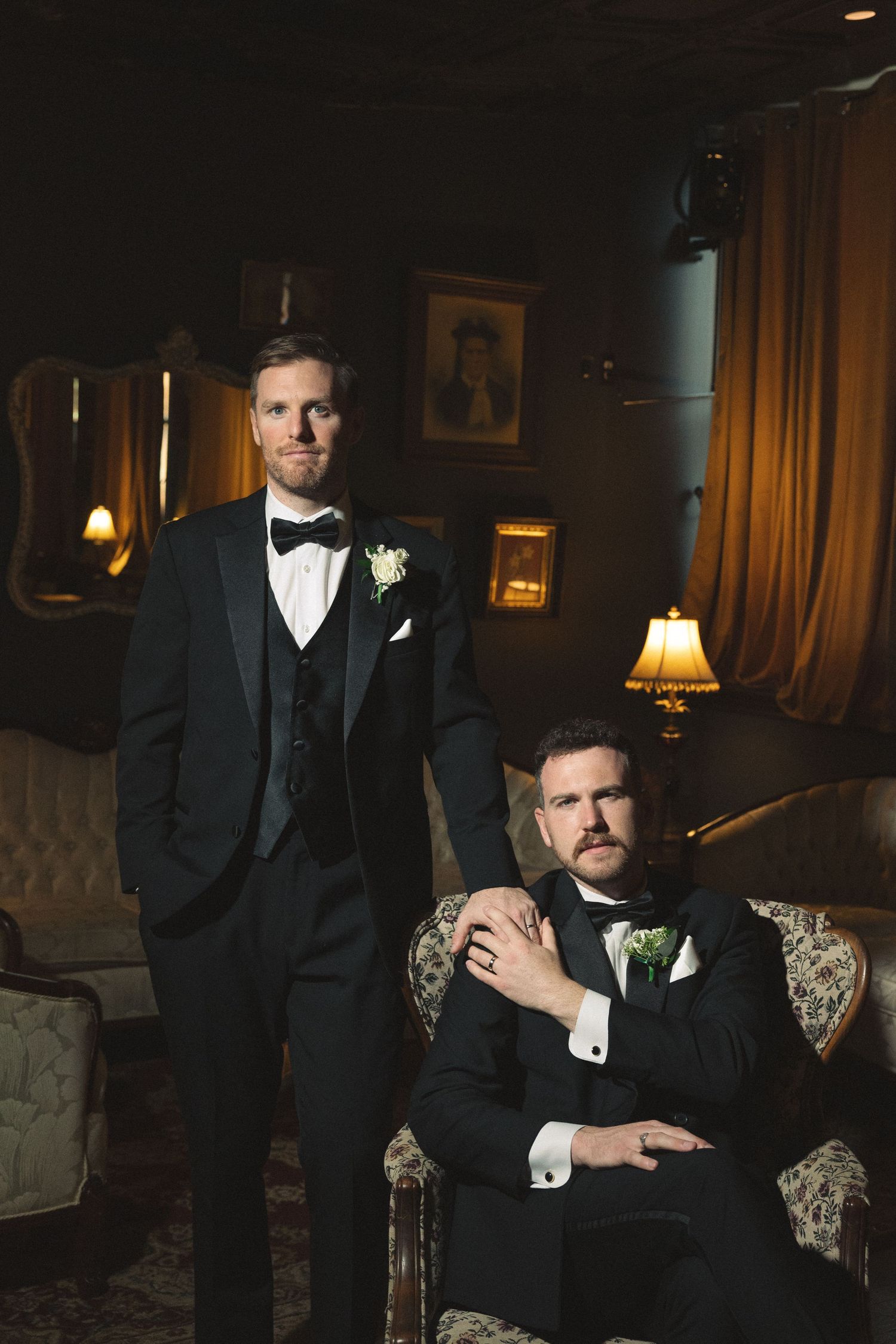 Two figures in formal attire sit elegantly on an antique sofa in moody lighting.