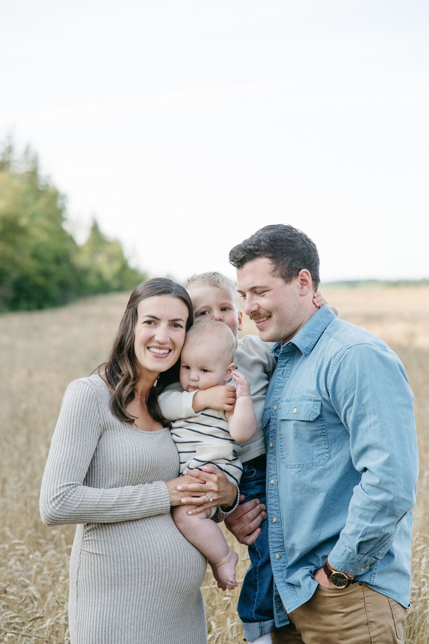 A happy pregnant woman and man hold two young children in a golden wheat field during a family photo session.