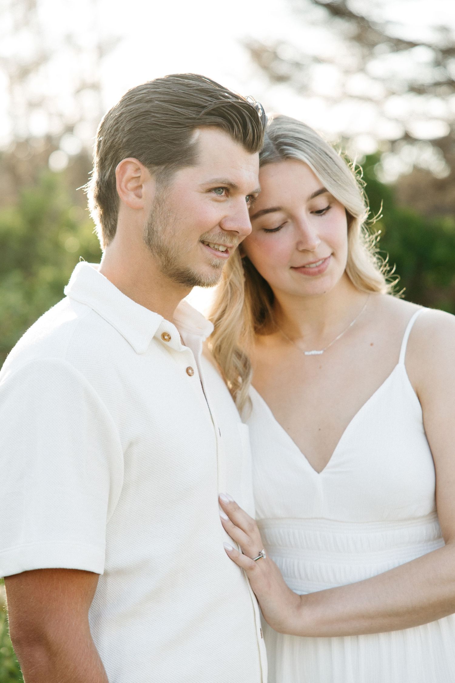 A couple in white outfits share a tender moment outdoors in golden hour light, the woman wearing a delicate necklace.