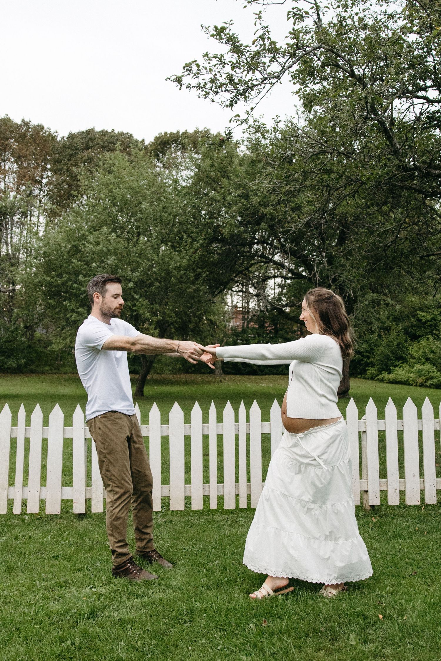 Pregnant couple holding hands and dancing together in a lush green backyard with a white picket fence.