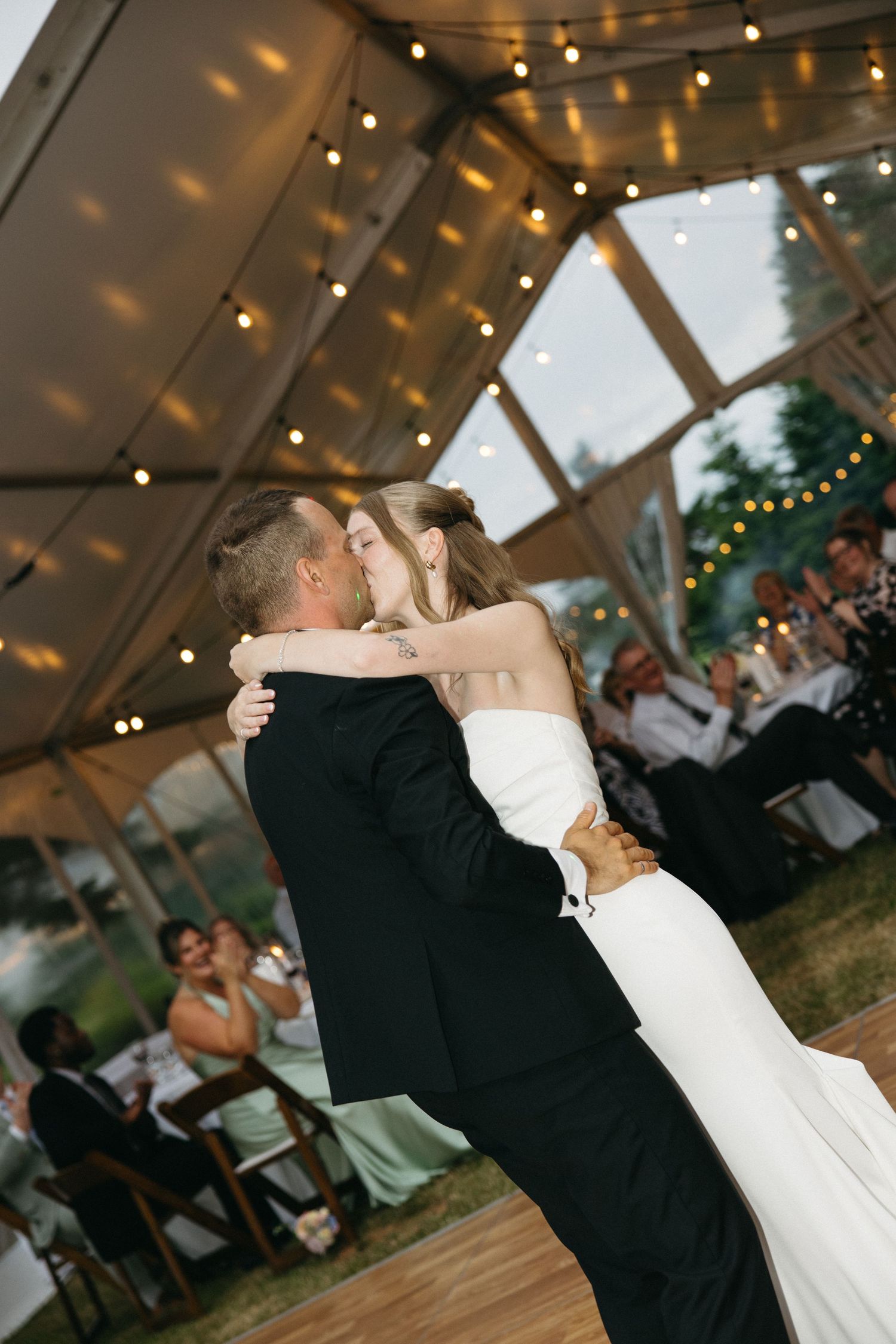 Bride and groom sharing romantic first dance kiss under string lights inside a clear wedding tent.