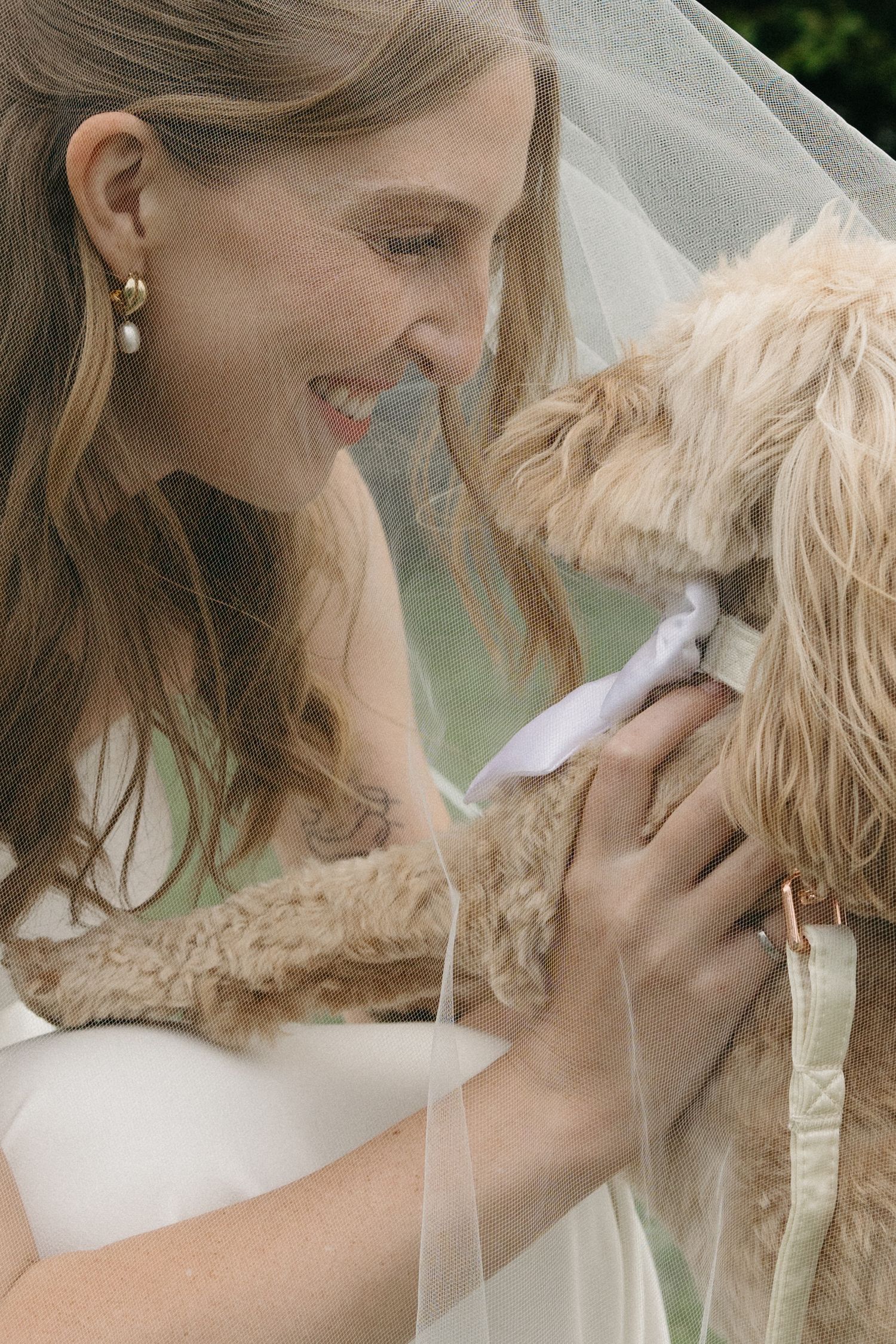 Smiling bride in veil tenderly holds fluffy dog wearing white bow tie at outdoor garden wedding.