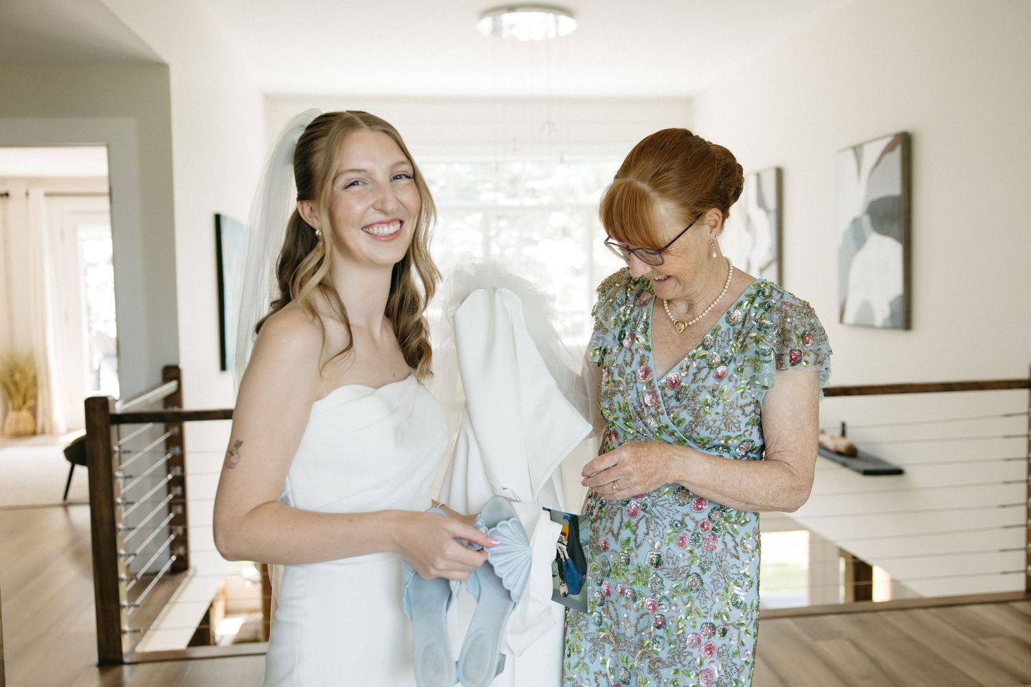 A smiling bride in a white strapless gown holds her veil while a woman in a floral dress assists her indoors.