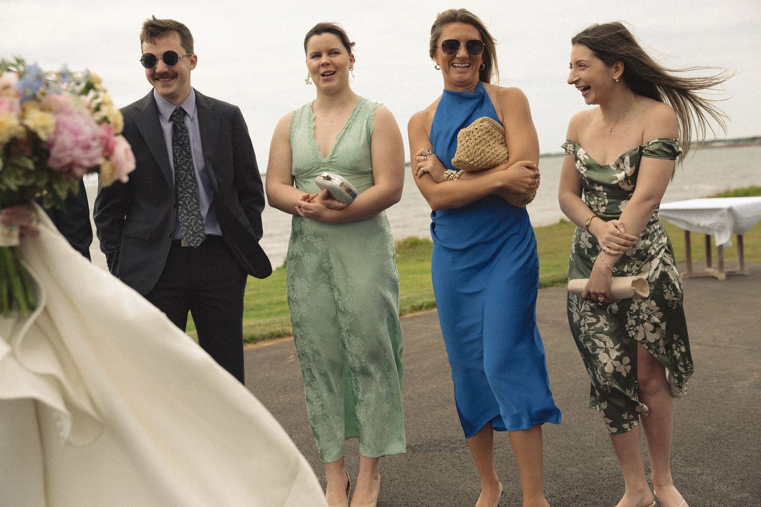 Four wedding guests laughing outdoors, wearing colorful dresses and suits near a floral bouquet on a windy day.