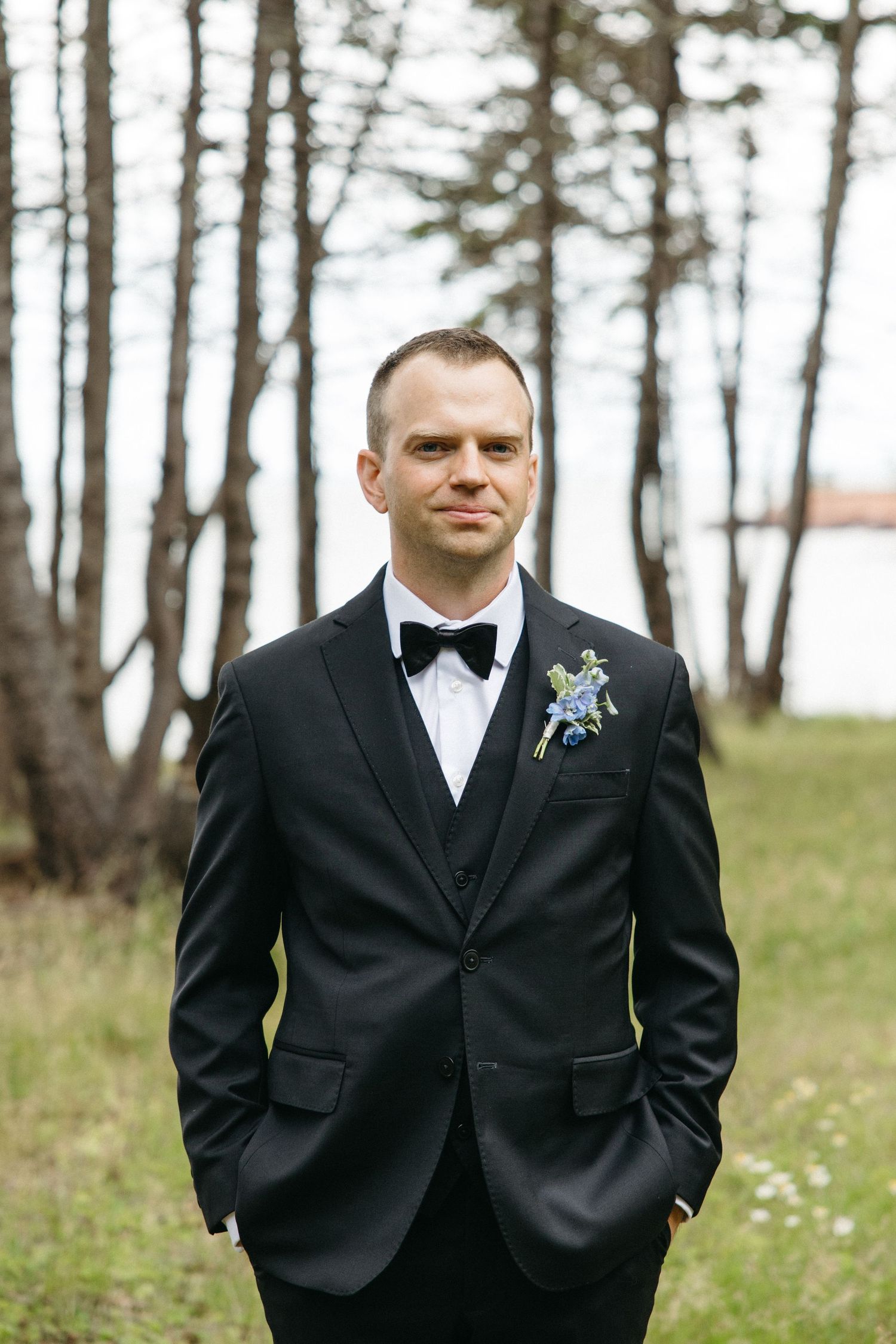 Dapper groom in black three-piece tuxedo with floral boutonniere poses outdoors near pine trees.