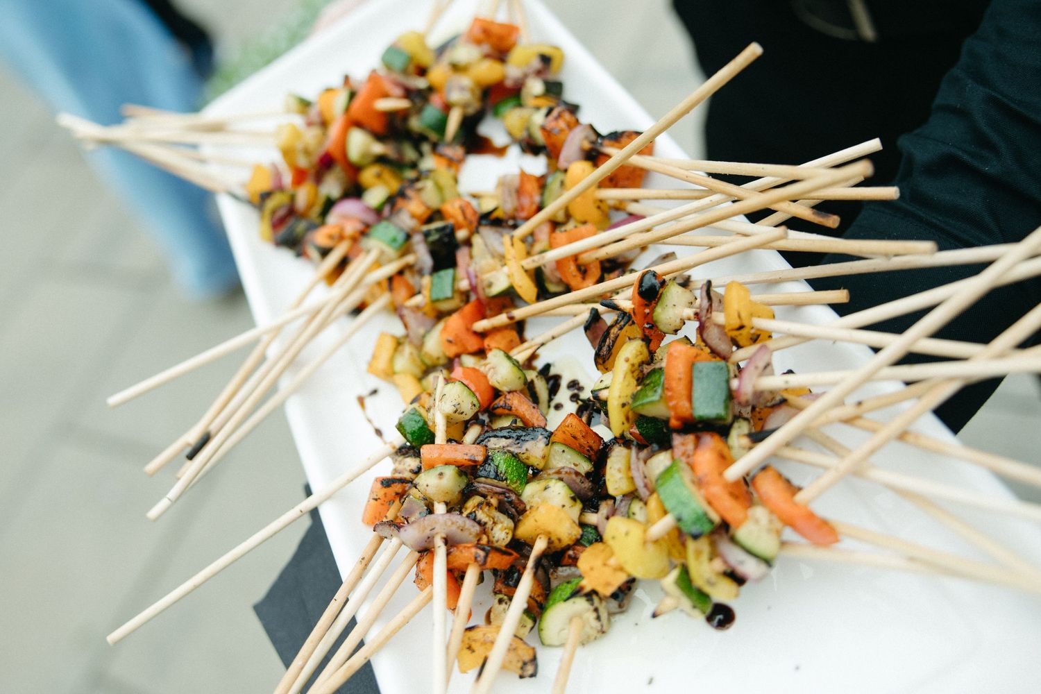 Colorful vegetable skewers on wooden sticks arranged on a white platter at an outdoor food event.