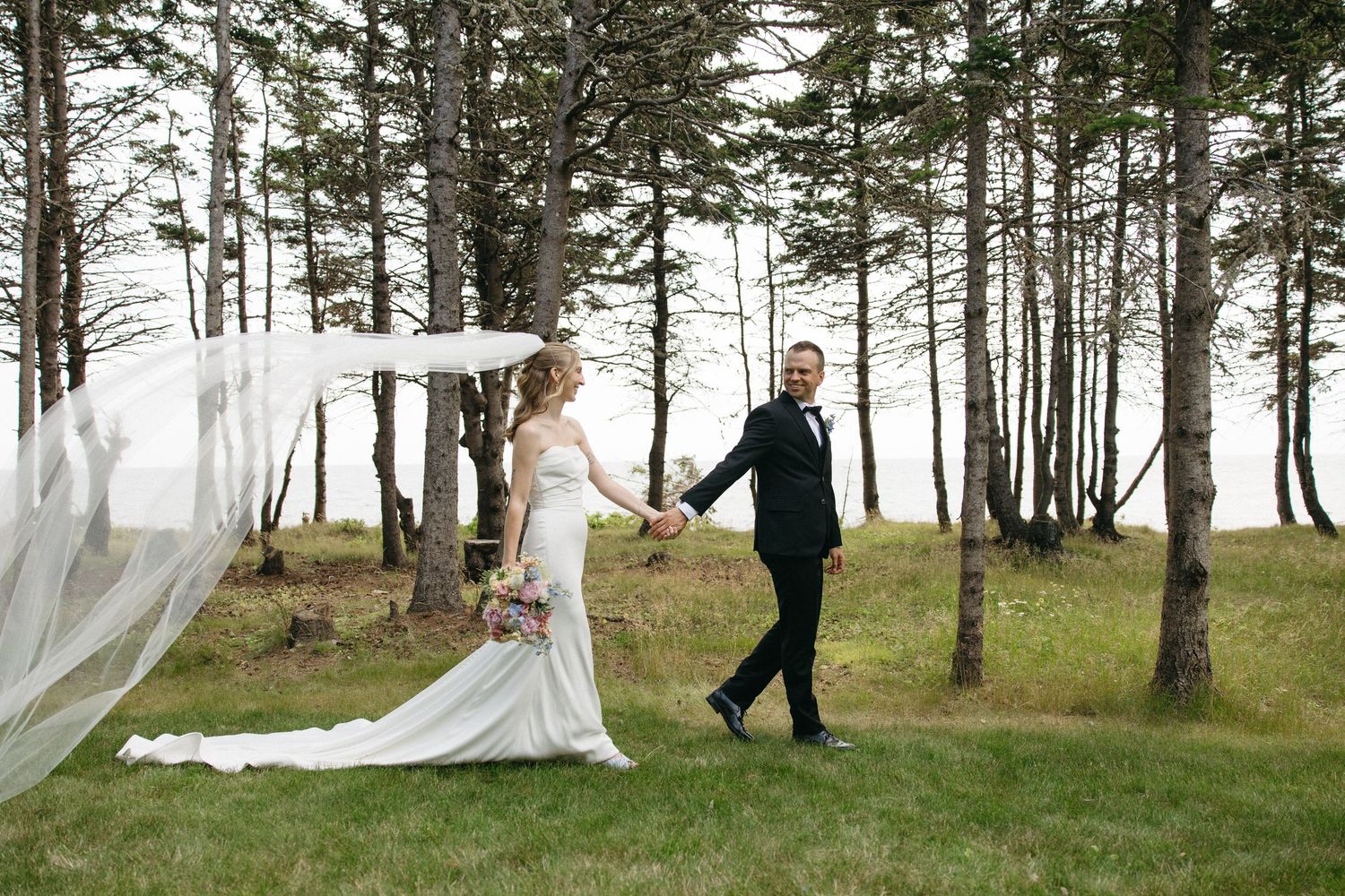 Bride and groom walk hand in hand through a pine tree grove, her long veil flowing in the wind.
