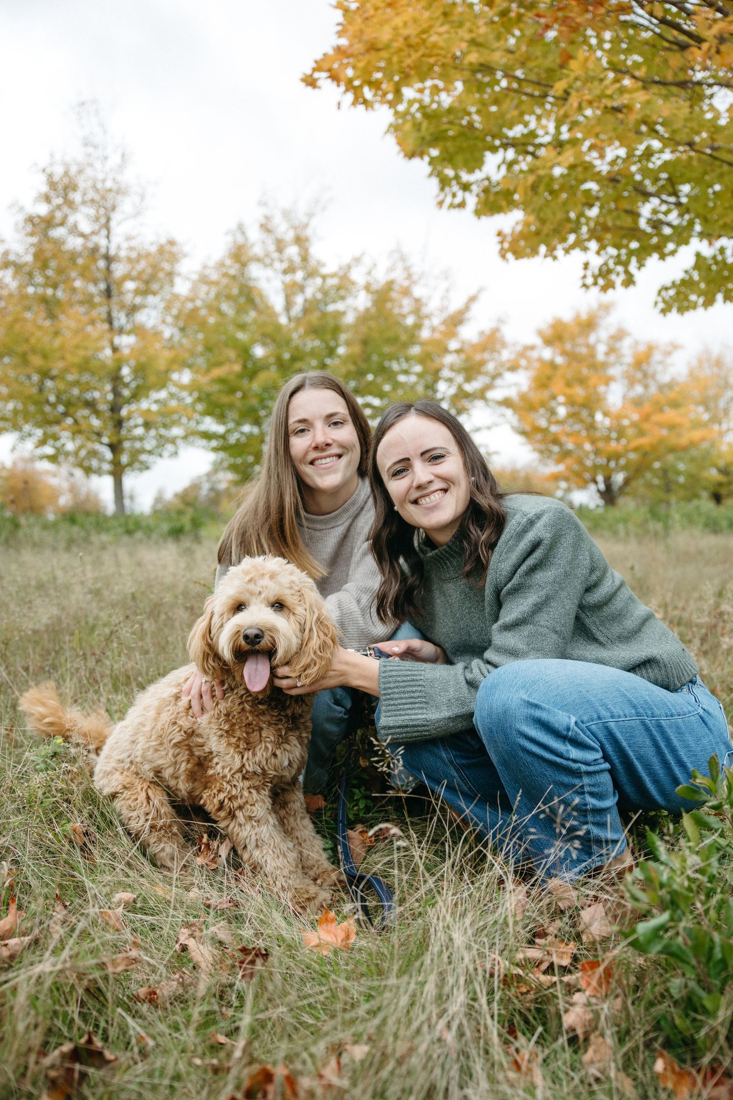 Two smiling women kneel in tall grass with a fluffy goldendoodle surrounded by colorful fall foliage.