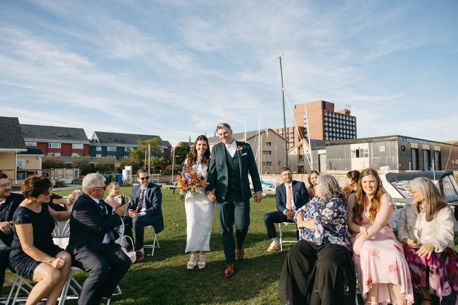 Bride holding a bouquet and groom in a green suit walk down the grass aisle past seated guests at an outdoor waterfront wedding ceremony.