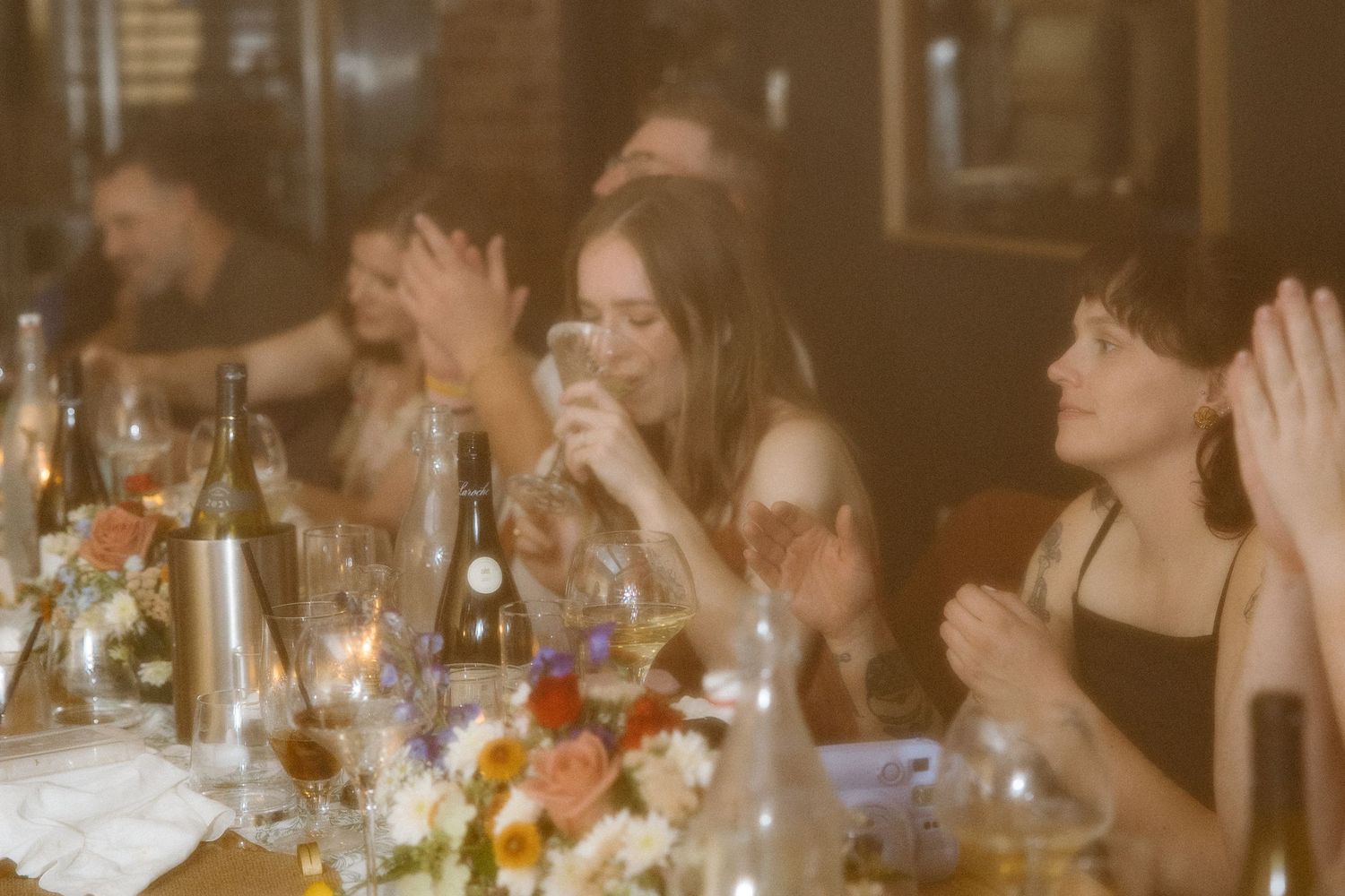 Guests laugh and raise glasses at a candlelit dinner table decorated with fresh flowers and wine bottles in a warm, cozy restaurant.