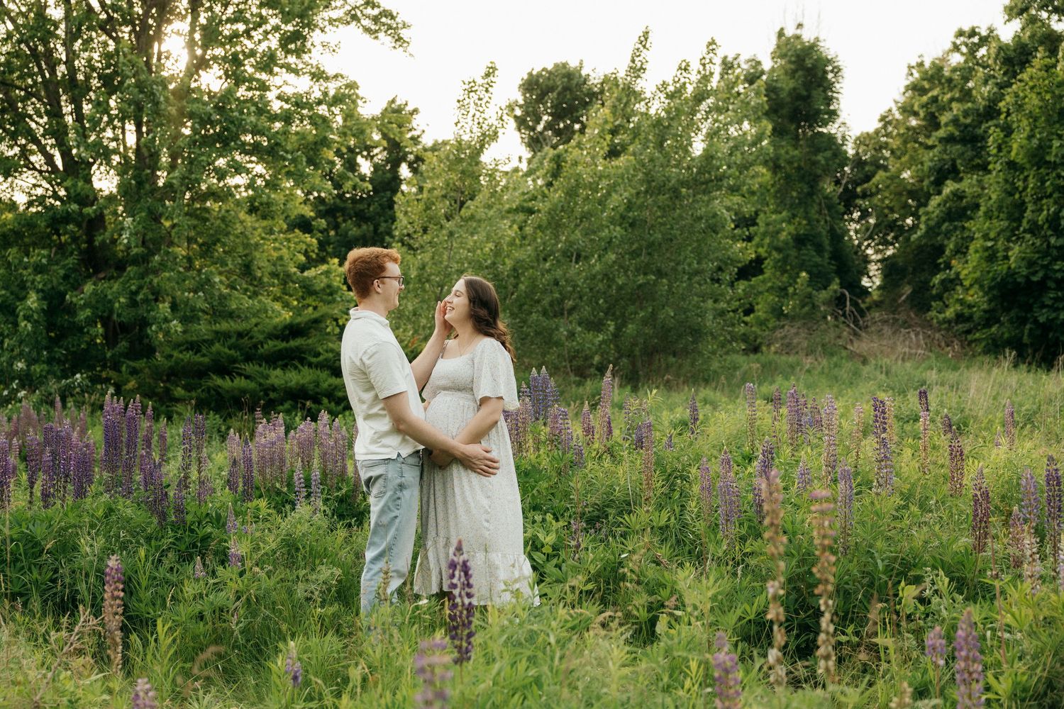 A couple embraces in a sunlit meadow with purple wildflowers blooming in the background.