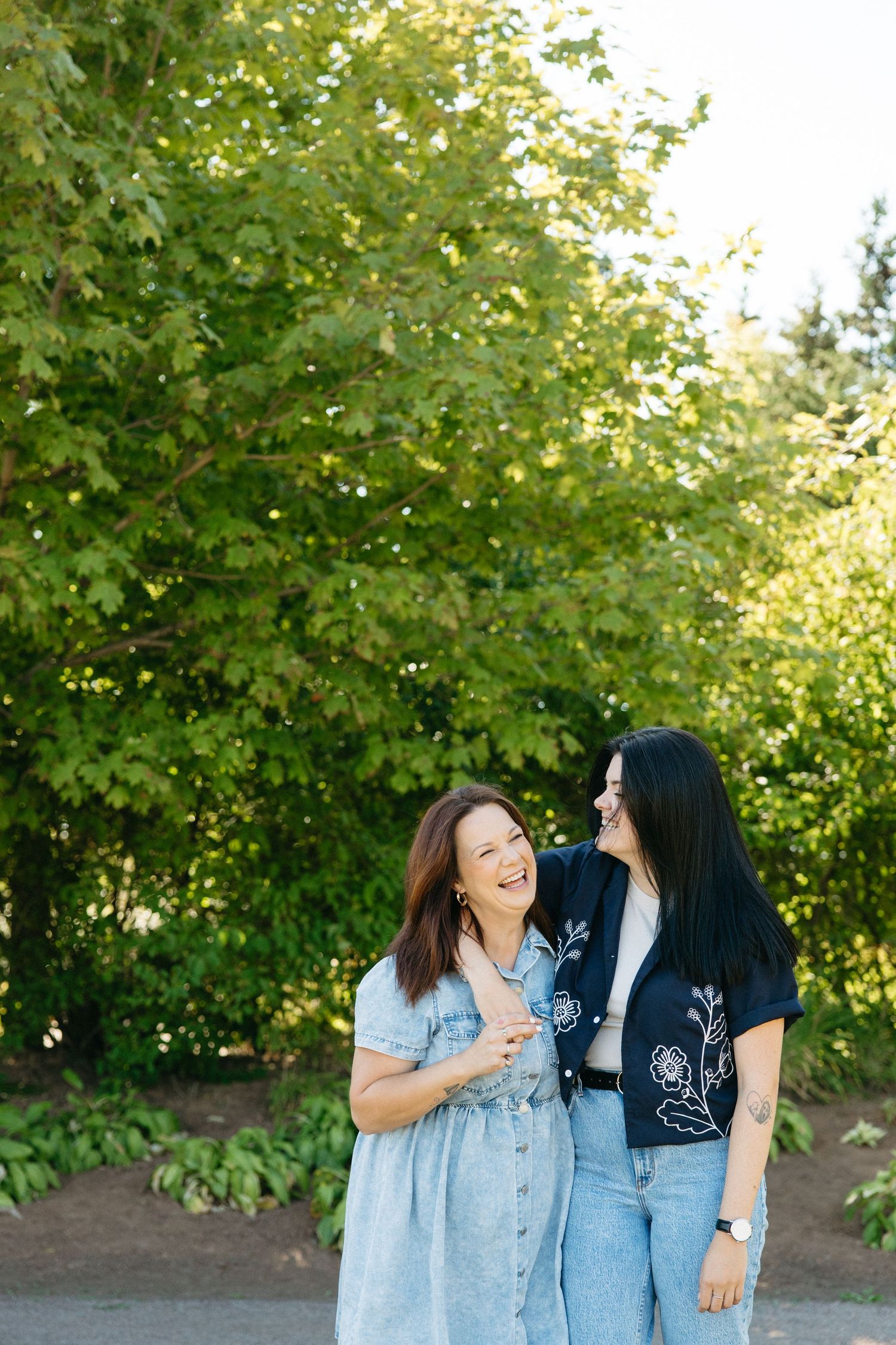 Two people in denim outfits laughing together outdoors with green foliage in background.