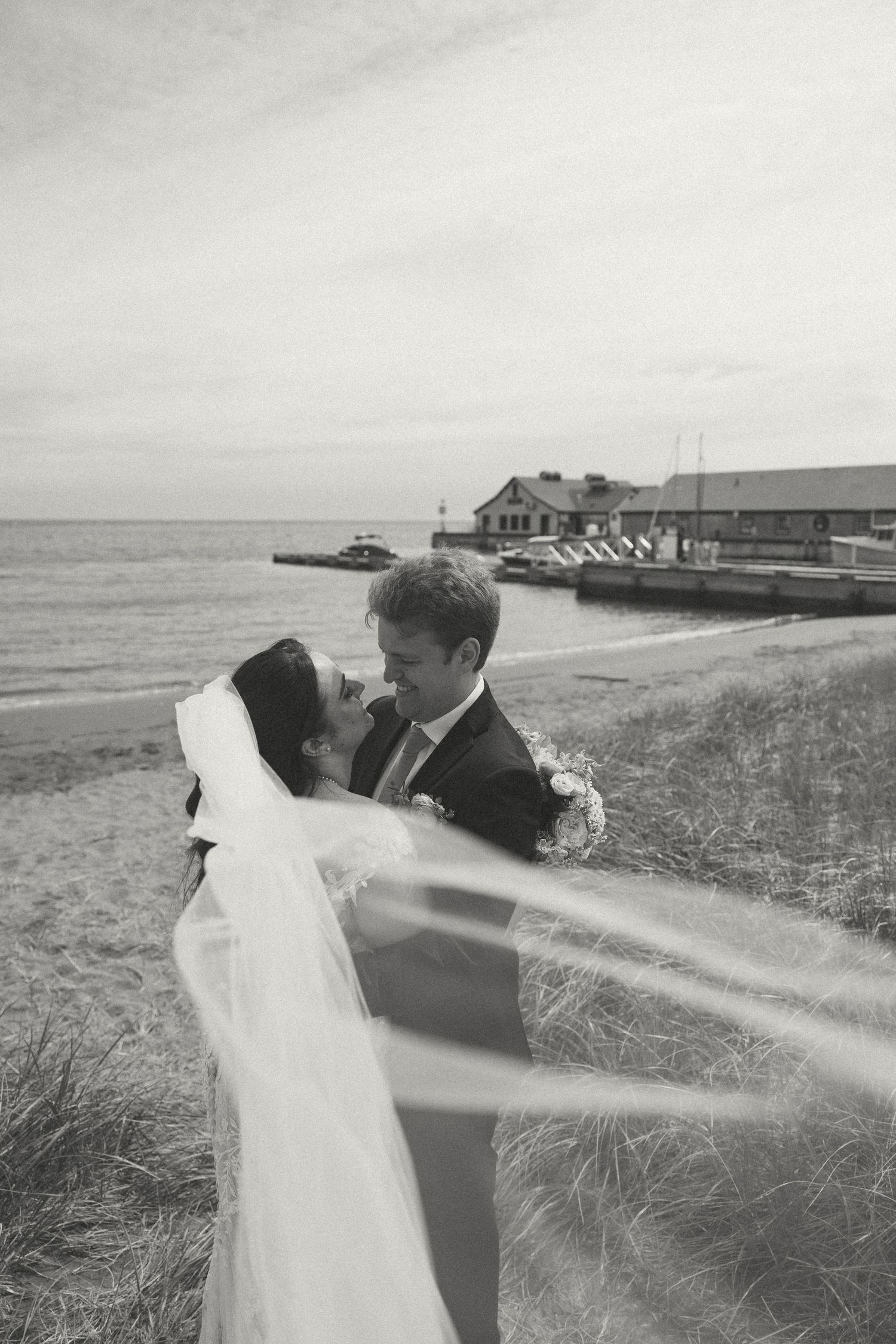 Black and white wedding photo of a couple embracing on a coastal path with dramatic veil movement.