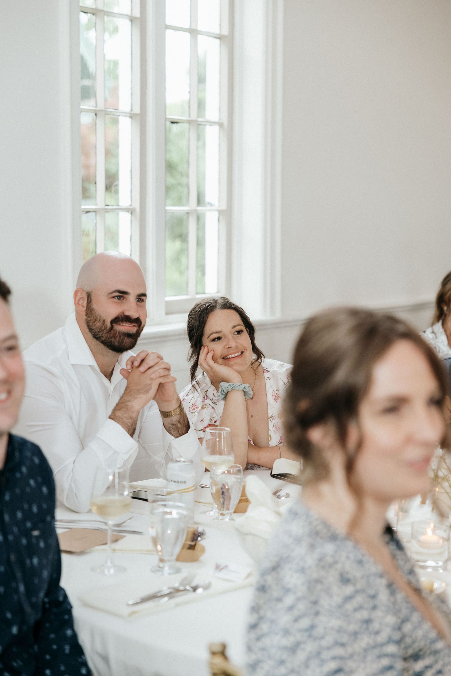 Wedding reception guests seated at white-clothed tables in bright indoor venue.