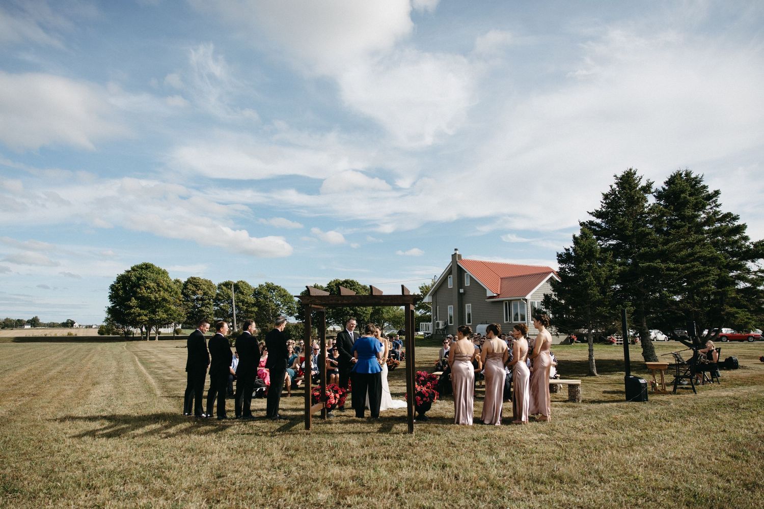 Wide landscape shot of wedding guests gathered for an outdoor ceremony near a coastal property.