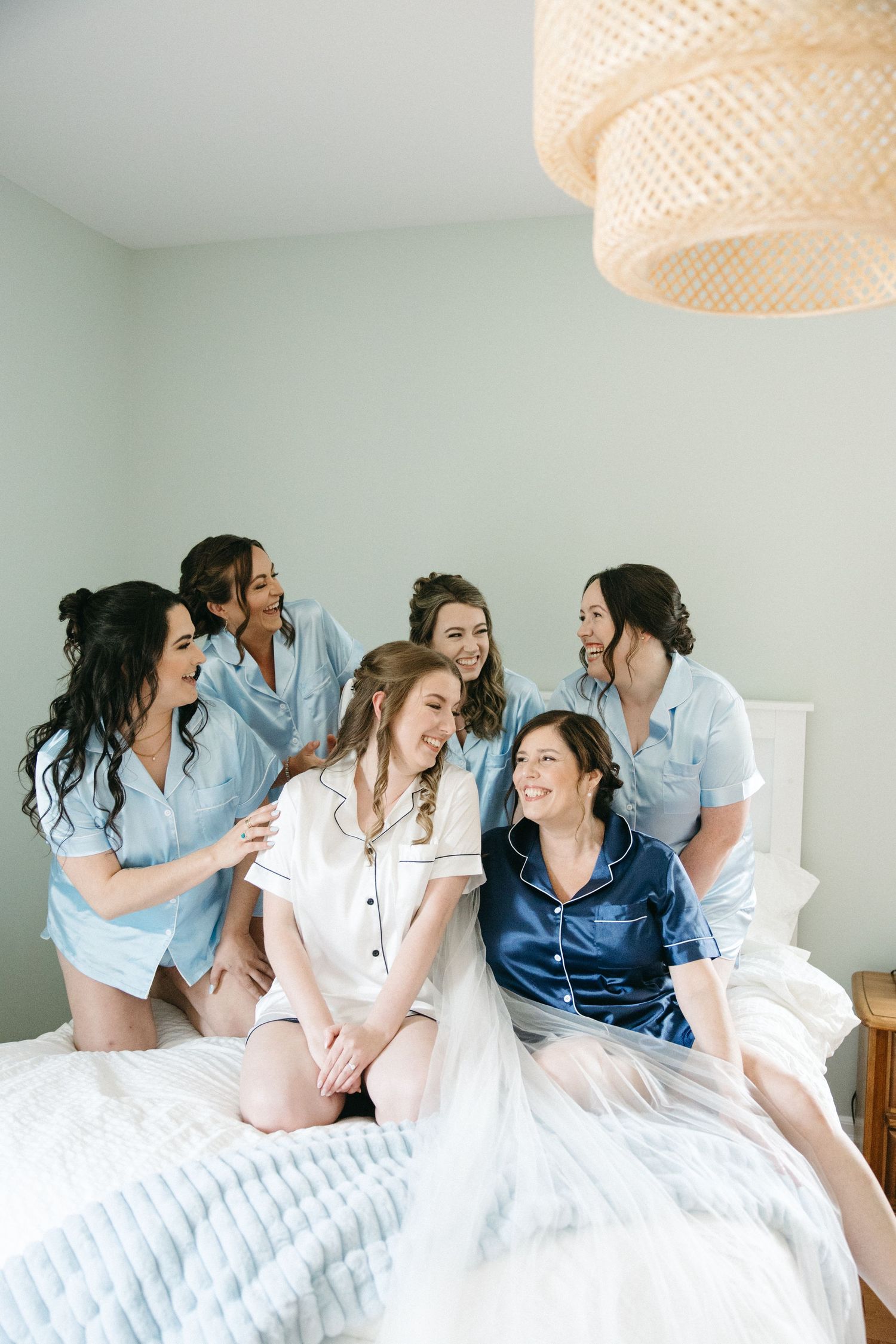 A group in matching blue and white sleepwear share joyful laughter while sitting on a bed under a woven pendant light.