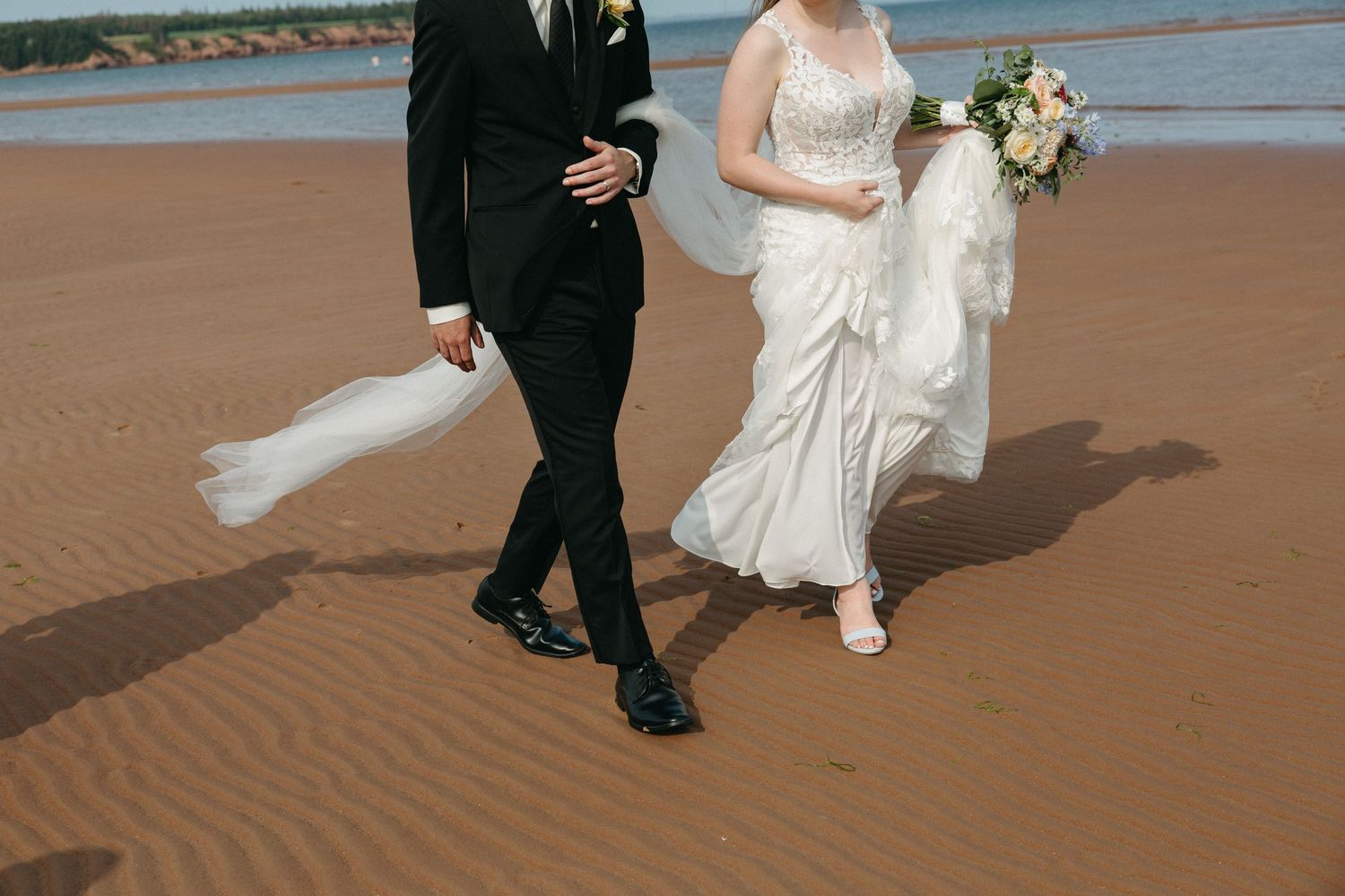 Couple walking along sandy beach shoreline in formal wedding attire.