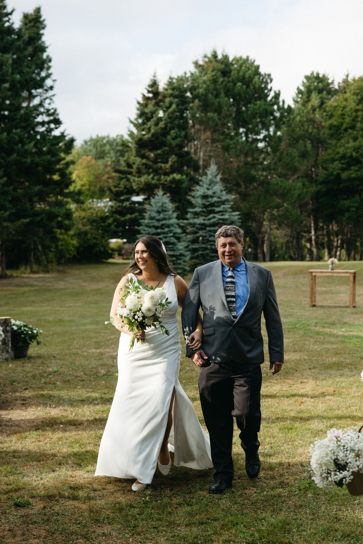 Wedding ceremony taking place on grassy lawn with evergreen trees in the background.