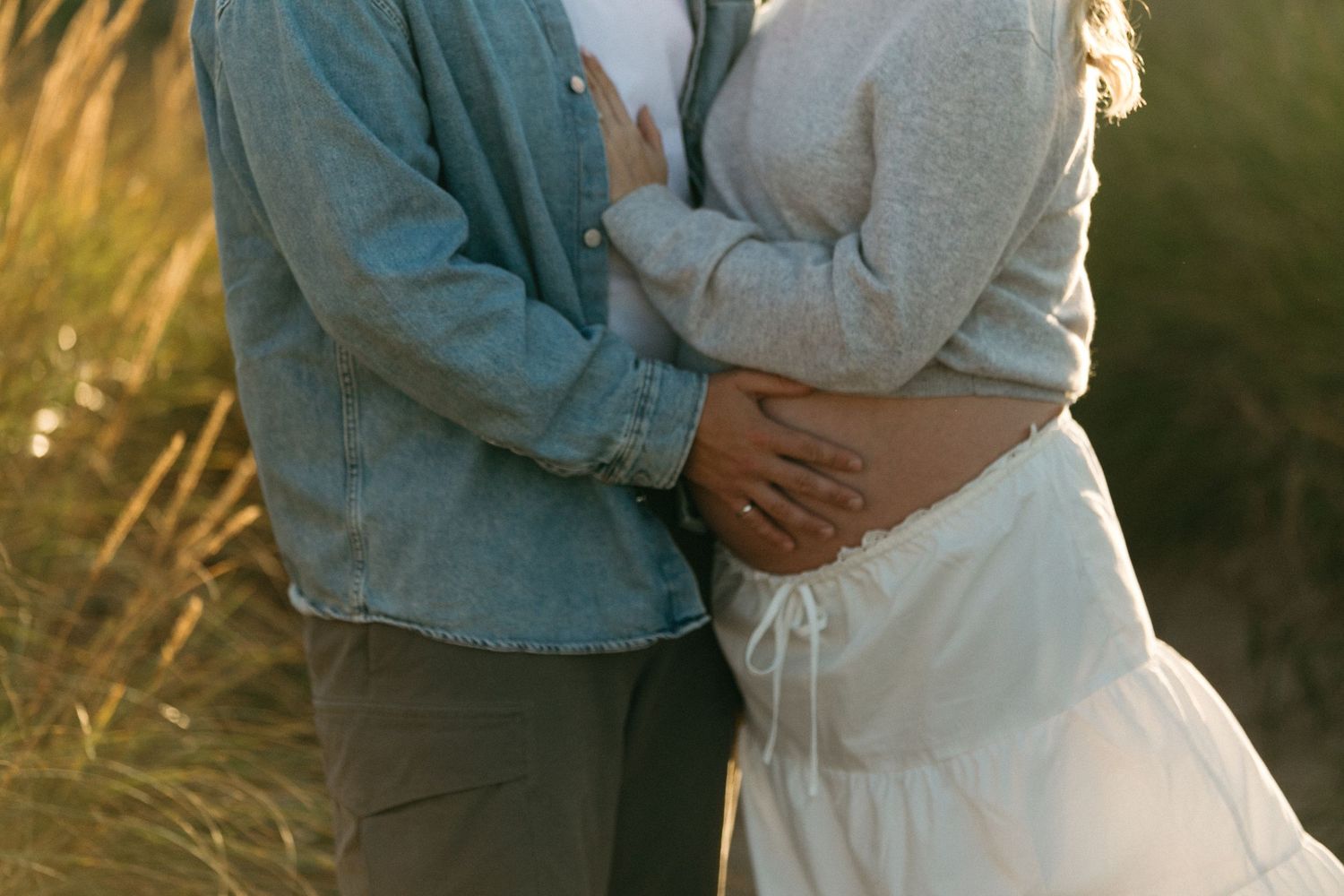 An intimate maternity moment captured in warm sunlight with coastal grasses.